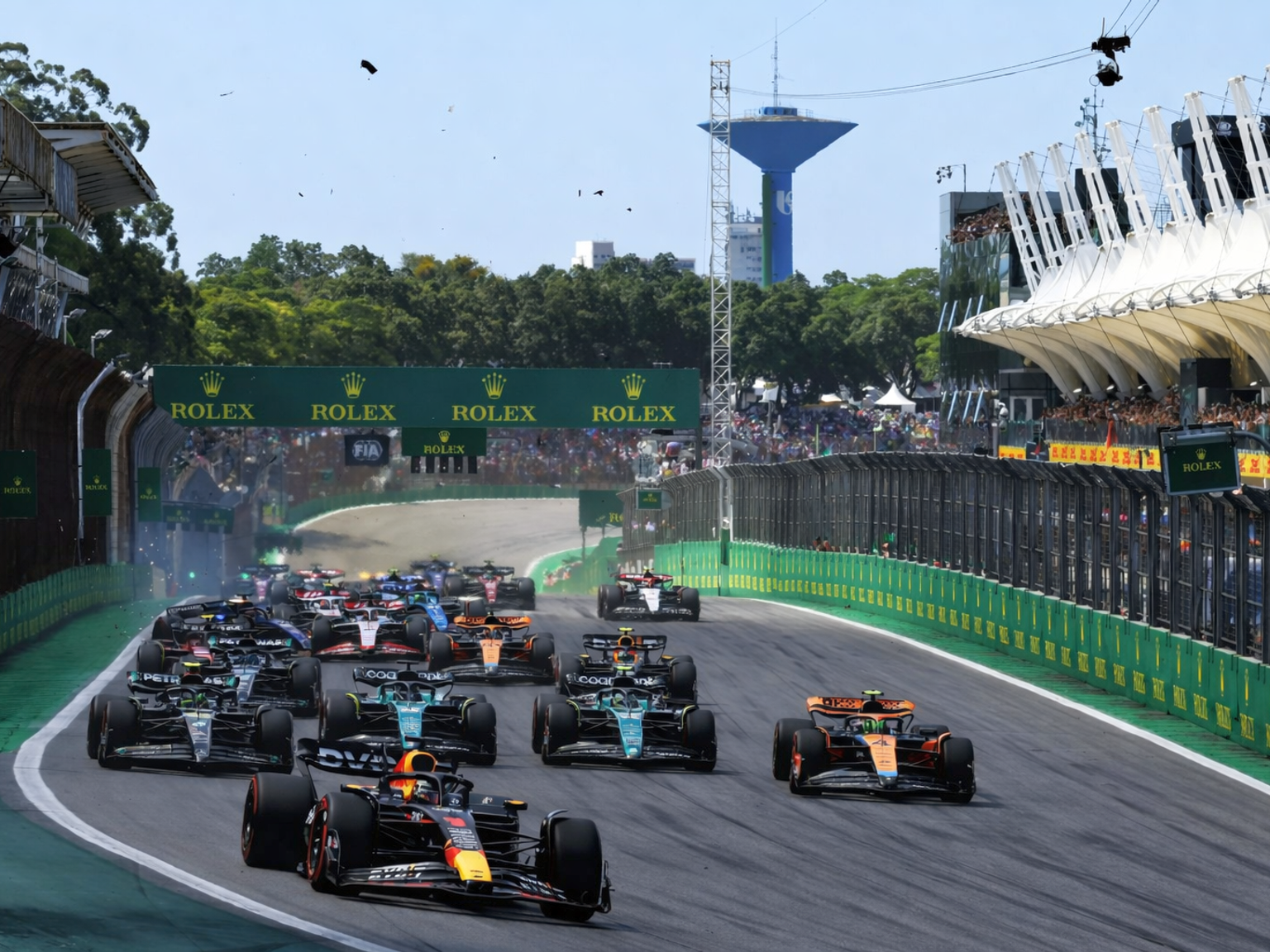 Numerous Formula 1 cars race on a track, passing grandstands filled with spectators.