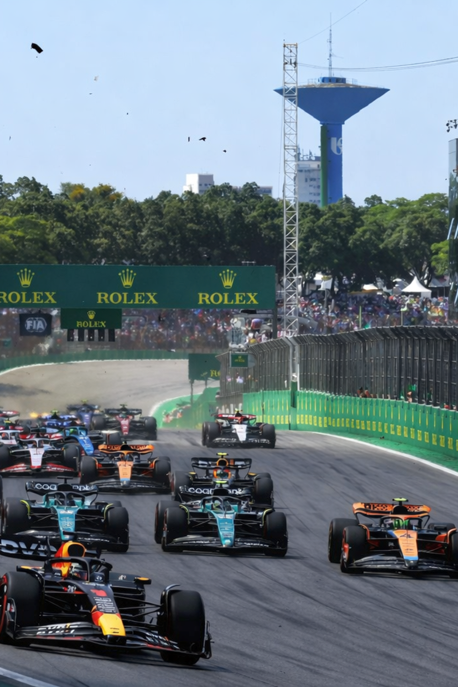 Numerous Formula 1 cars race on a track, passing grandstands filled with spectators.