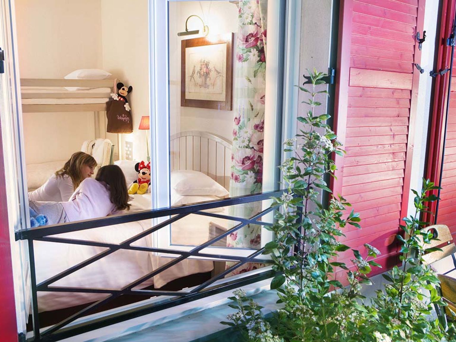 Två flickor pratar på en säng i ett sovrum med våningssängar och Disney-leksaker, sett genom ett fönster med röda fönsterluckor. Hotel Campanile Val de France vid Disneyland® Paris