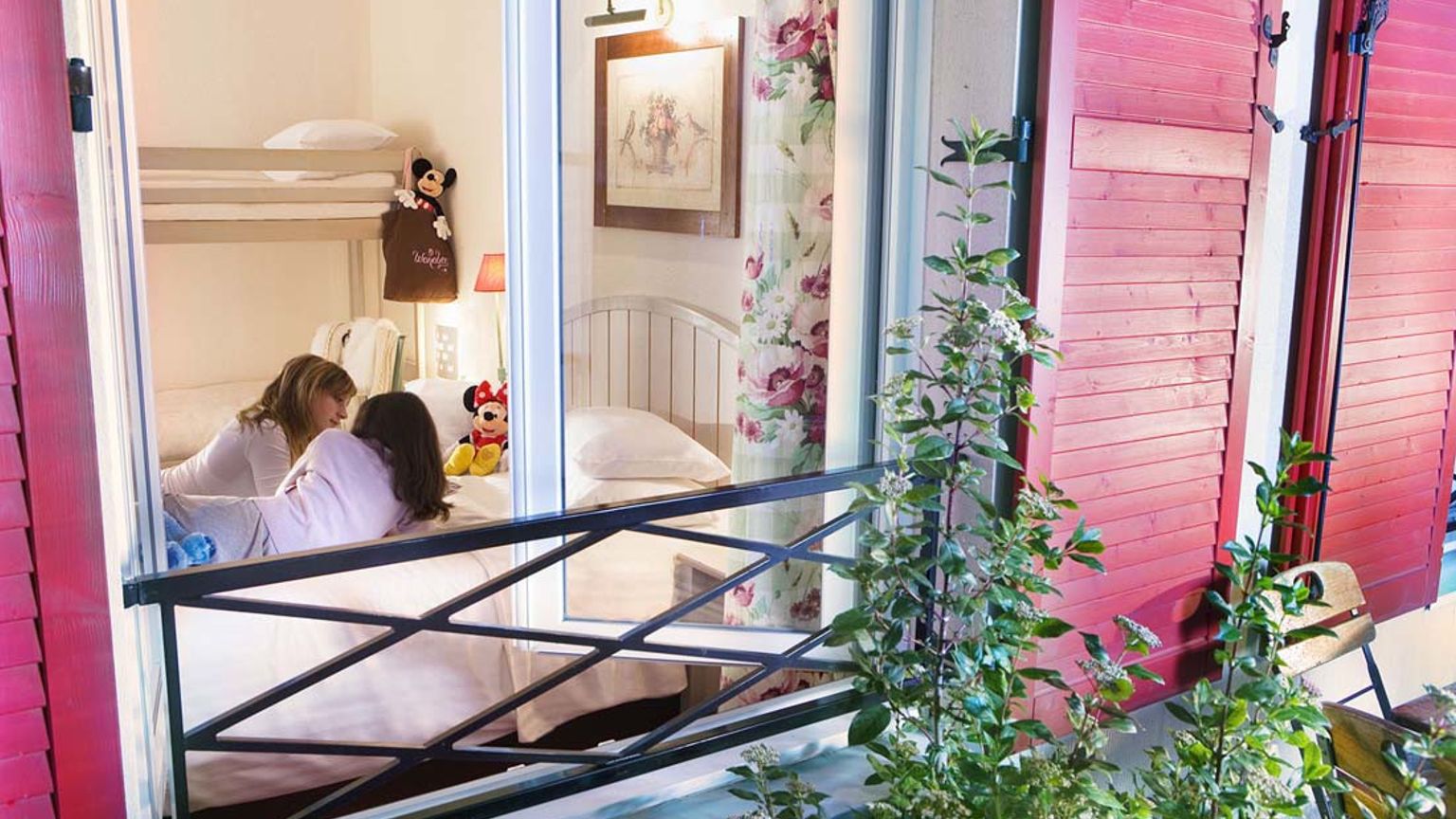Två flickor pratar på en säng i ett sovrum med våningssängar och Disney-leksaker, sett genom ett fönster med röda fönsterluckor. Hotel Campanile Val de France vid Disneyland® Paris