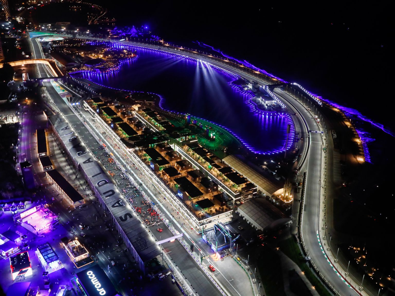Luftaufnahme bei Nacht von einer hell erleuchteten Formel-1-Rennstrecke und Einrichtungen neben einem Gewässer, das mit violettem Licht beleuchtet ist.