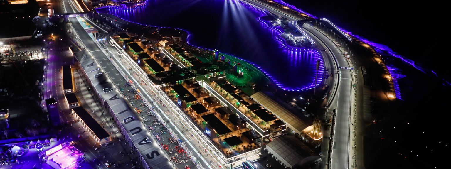 Luftaufnahme bei Nacht von einer hell erleuchteten Formel-1-Rennstrecke und Einrichtungen neben einem Gewässer, das mit violettem Licht beleuchtet ist.
