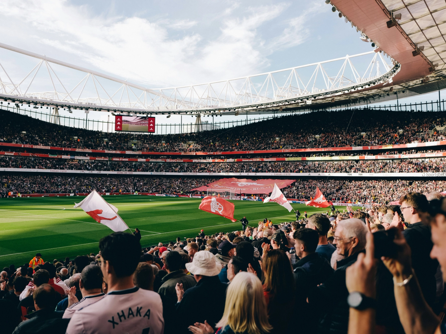 Packed football stadium with fans holding flags, overlooking a bright green pitch.