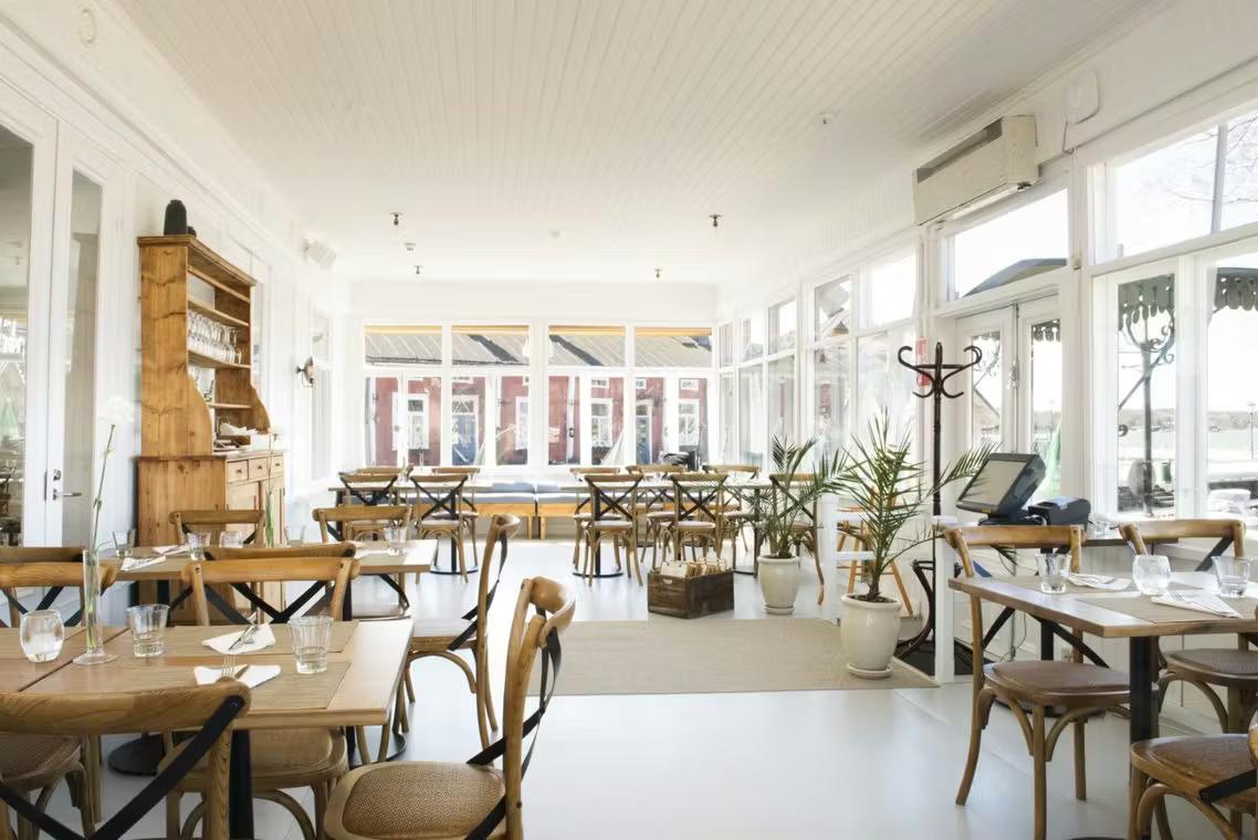 Lys, tom restaurant med hvidt interiør, lyse træborde og stole samt store vinduer.