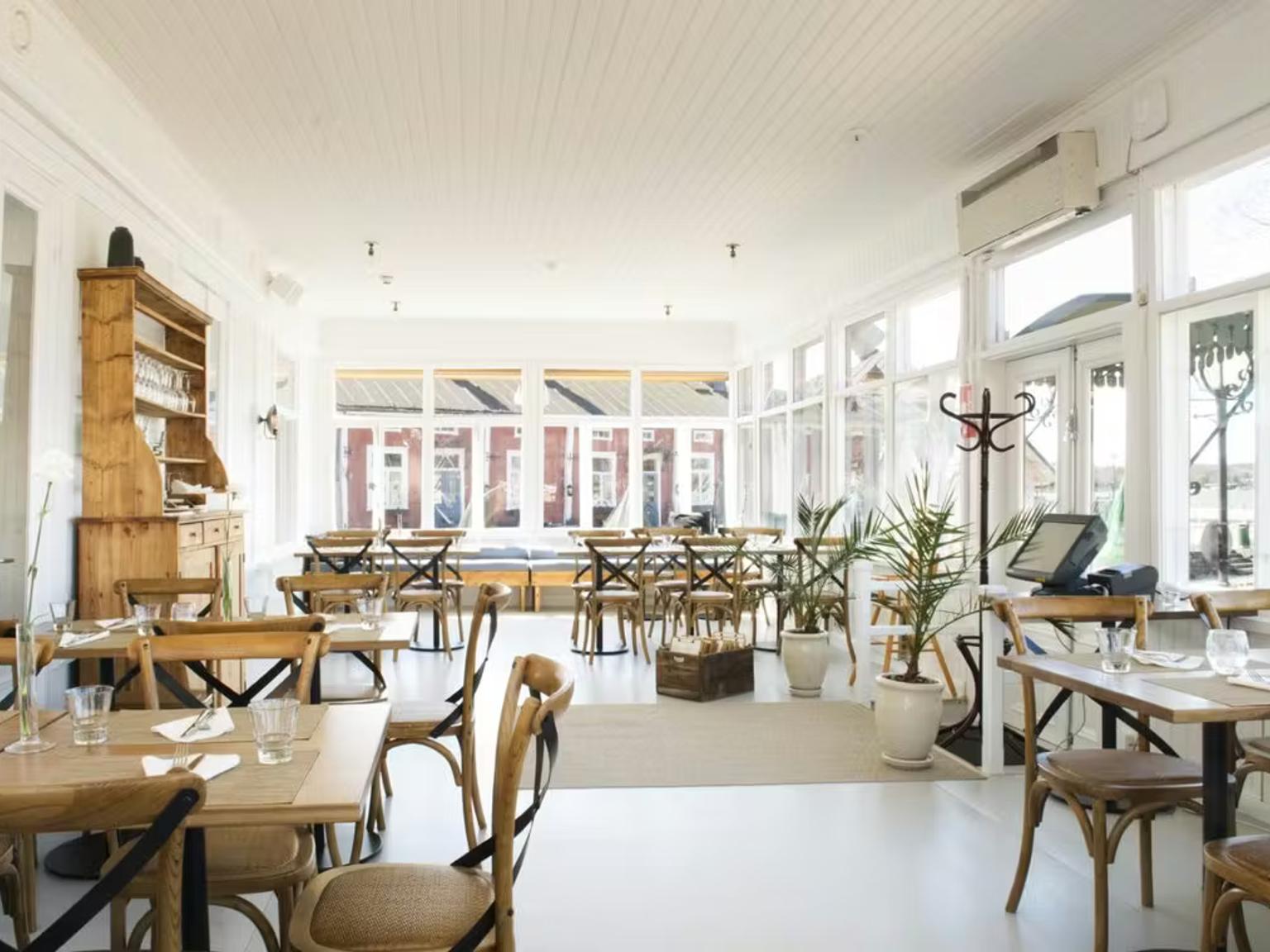 Lys, tom restaurant med hvidt interiør, lyse træborde og stole samt store vinduer.