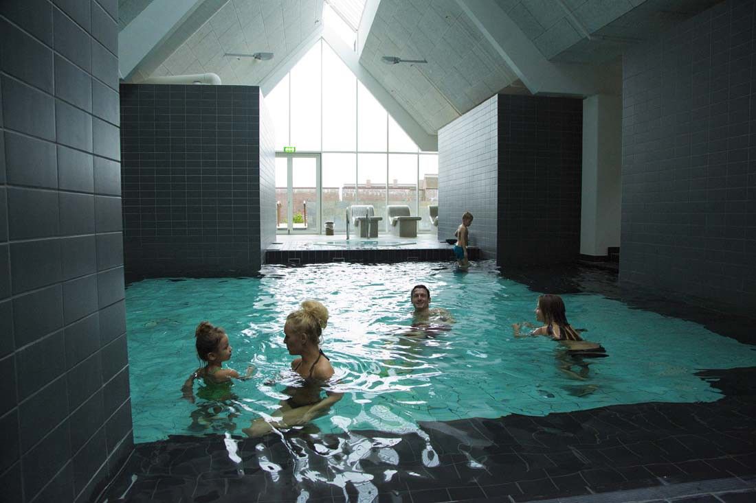 En familj på fem simmar i en modern inomhuspool med mörka kakelplattor, ett högt vinklat tak och stora fönster.