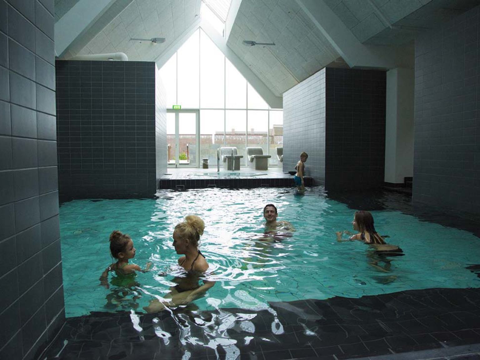 En familj på fem simmar i en modern inomhuspool med mörka kakelplattor, ett högt vinklat tak och stora fönster.