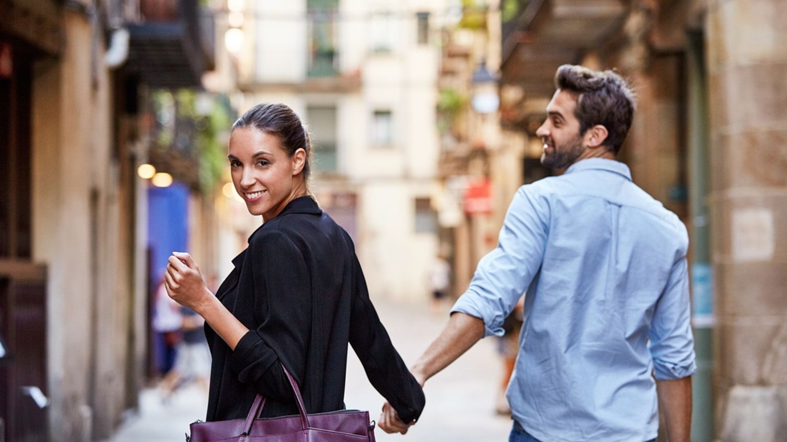 A smiling couple holding hands walks down a narrow street, the woman looking back at the camera.