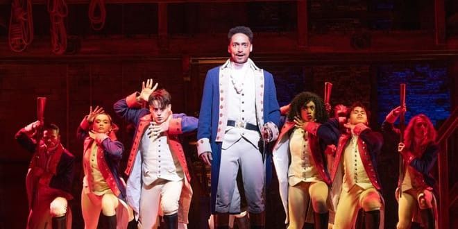 En scen från Hamilton, med en huvudskådespelare i blå rock och en ensemble i historiska kostymer som intar dramatiska poser på en rödlyst scen. Musikaler - Hamilton - London