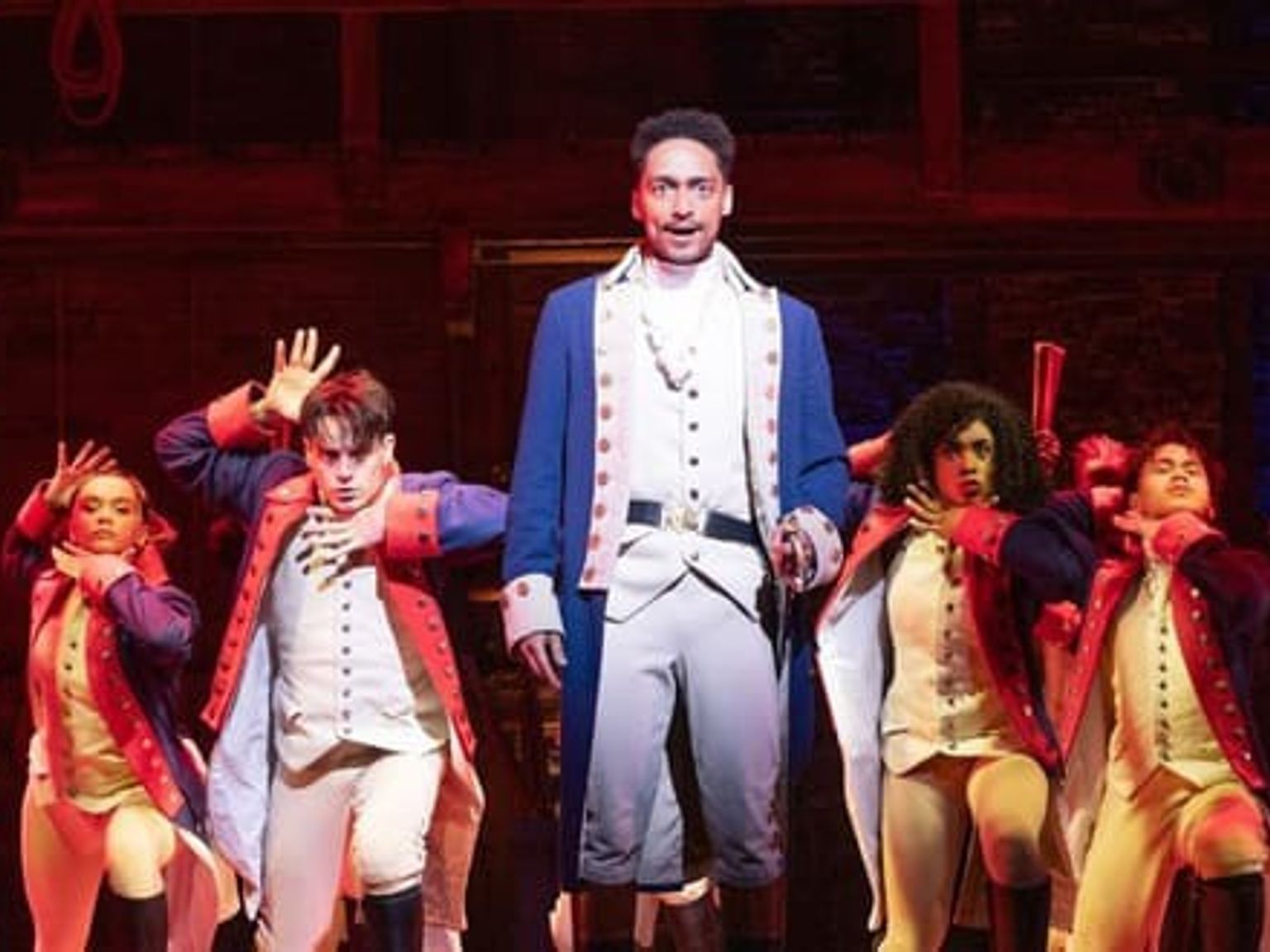 En scen från Hamilton, med en huvudskådespelare i blå rock och en ensemble i historiska kostymer som intar dramatiska poser på en rödlyst scen. Musikaler - Hamilton - London