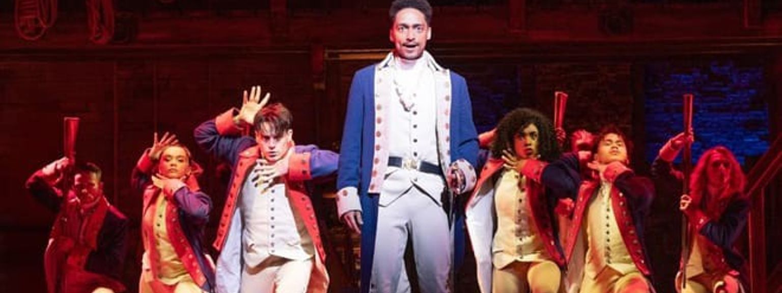 En scen från Hamilton, med en huvudskådespelare i blå rock och en ensemble i historiska kostymer som intar dramatiska poser på en rödlyst scen. Musikaler - Hamilton - London