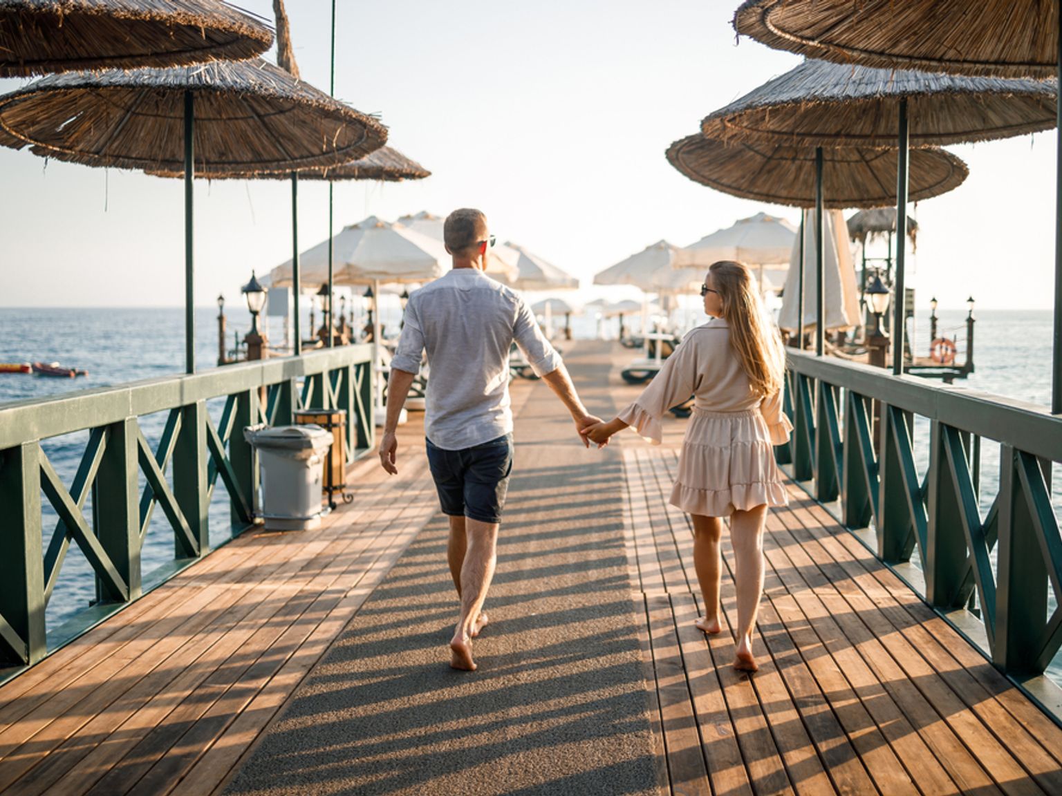 Ett par går hand i hand nerför en träpir som kantas av parasoller av halm.