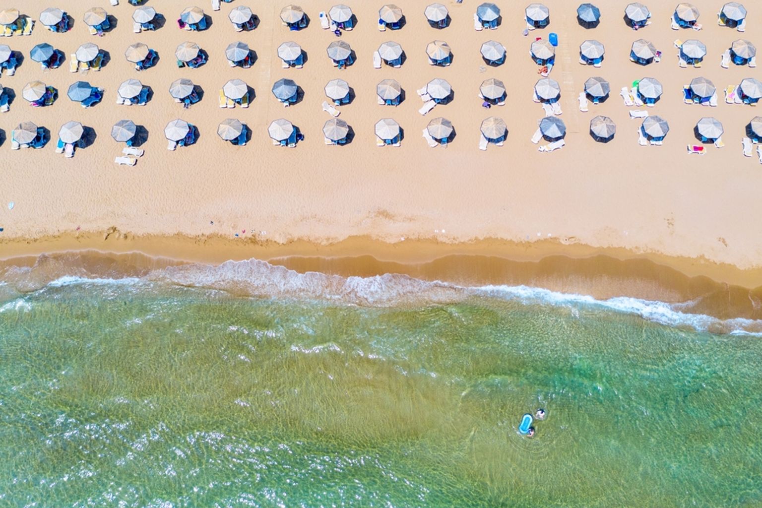 Flygfoto över en sandstrand med rader av parasoller och klart havsvatten där en person paddlar.