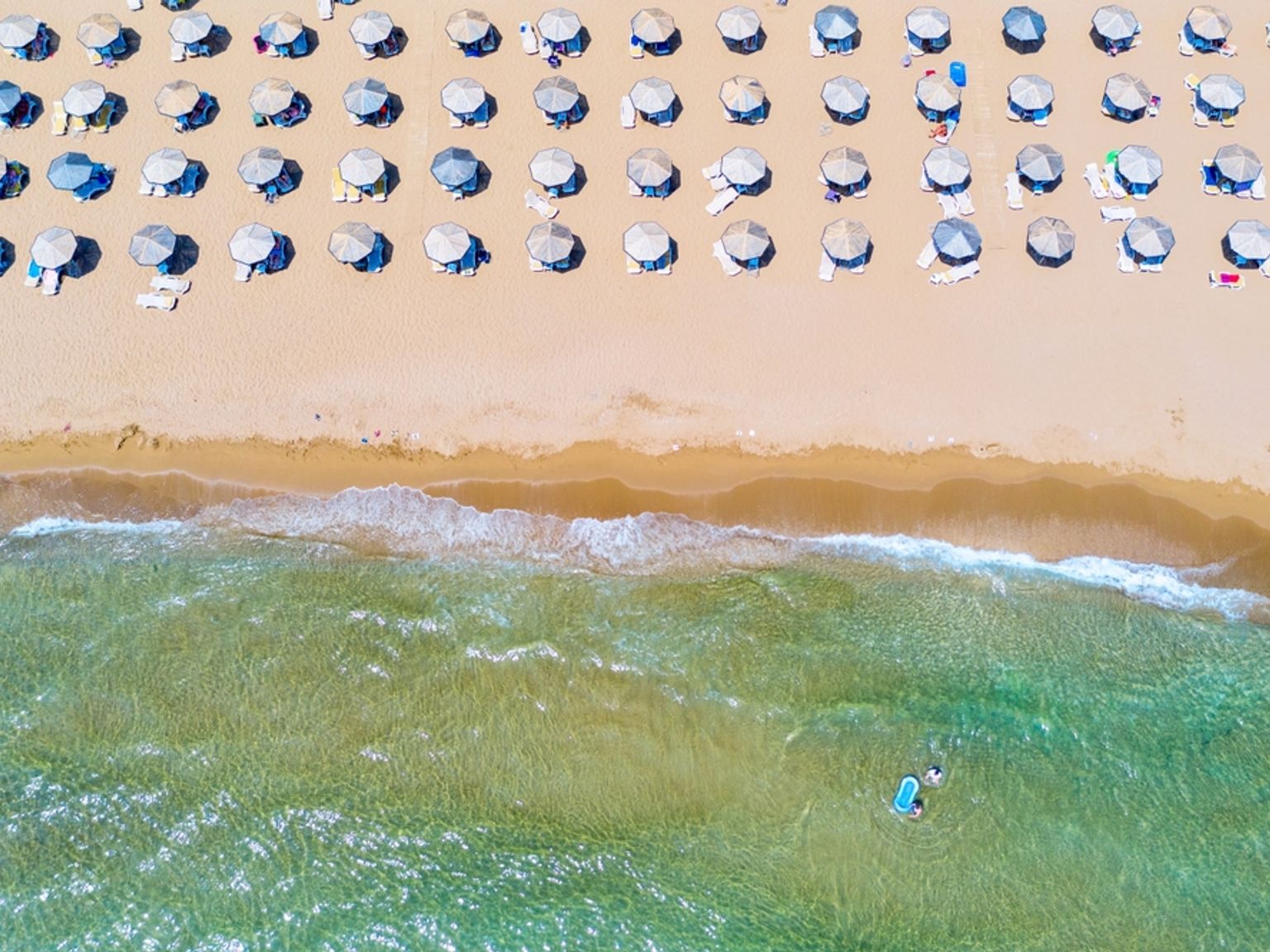 Flygfoto över en sandstrand med rader av parasoller och klart havsvatten där en person paddlar.