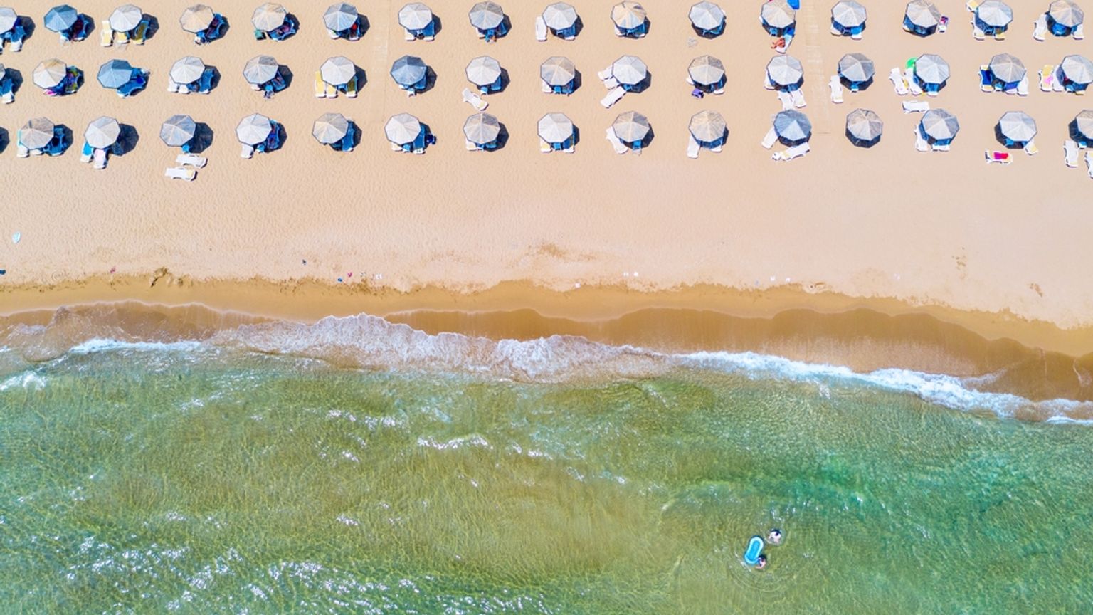 Flygfoto över en sandstrand med rader av parasoller och klart havsvatten där en person paddlar.