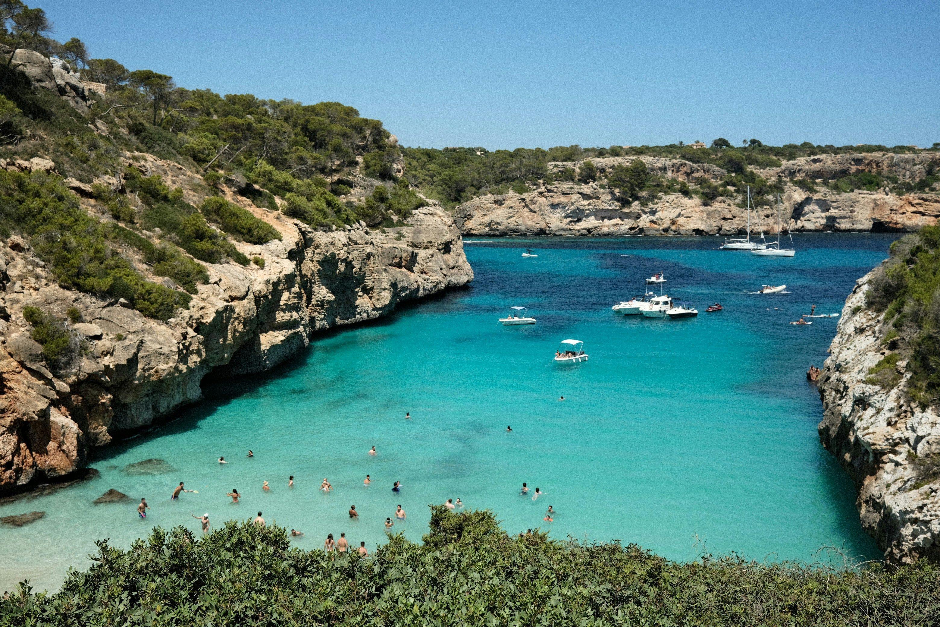 Una cala de agua turquesa cristalina con personas nadando y barcos, rodeada de acantilados rocosos cubiertos de árboles. Mallorca, España