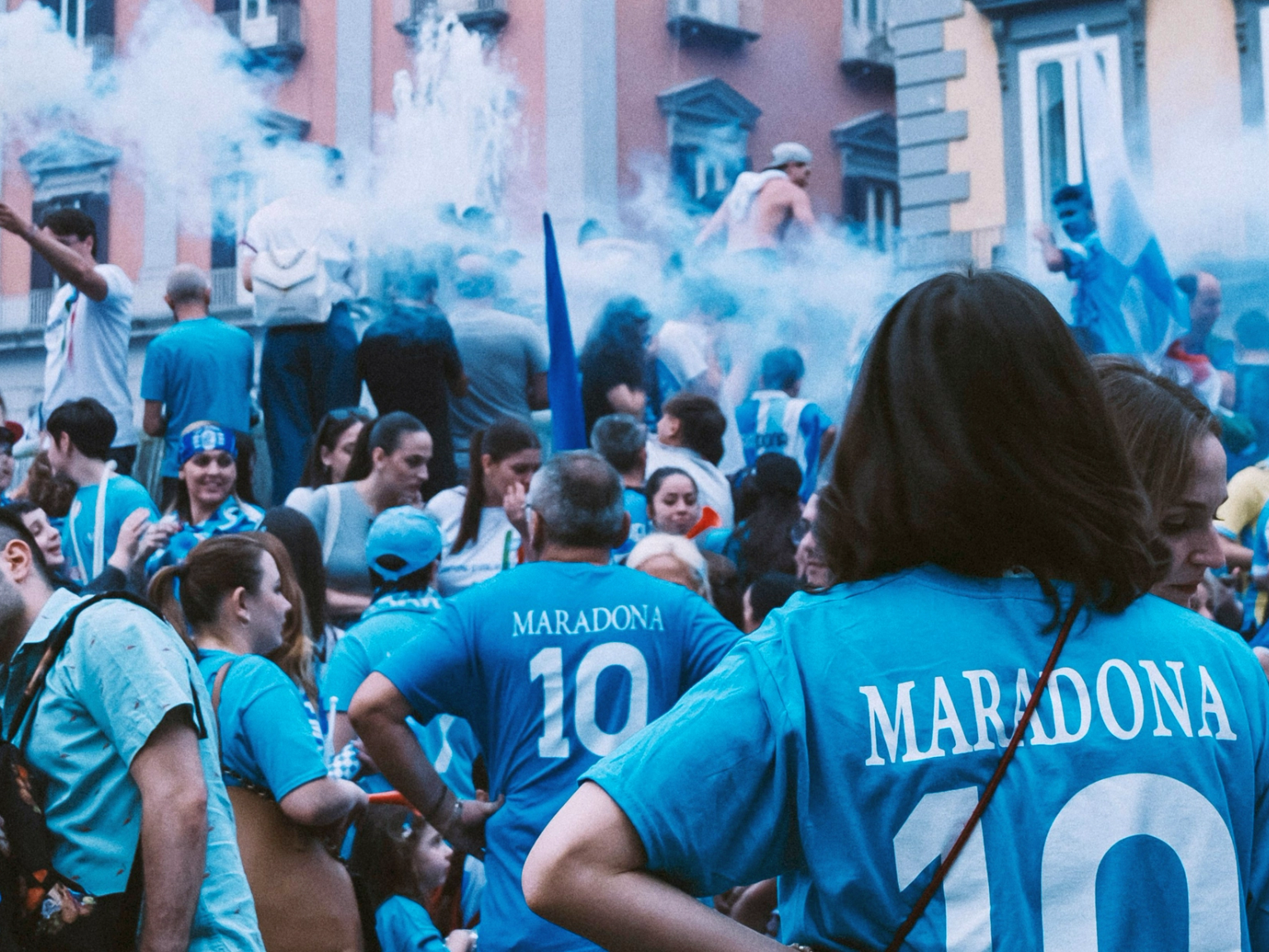 Uma grande multidão de pessoas, muitas com camisetas azuis "Maradona 10", reunidas com fumaça azul no ar.