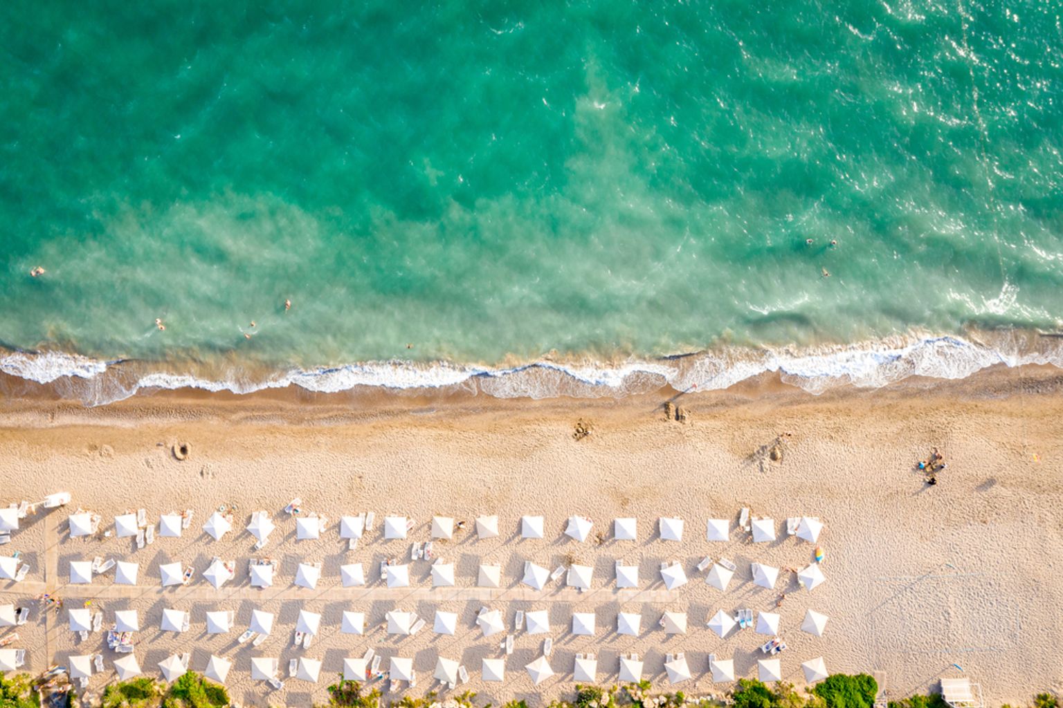 Vue aérienne d'une plage avec de l'eau turquoise, un rivage sablonneux et de nombreux parasols et transats blancs. Belek, Turquie