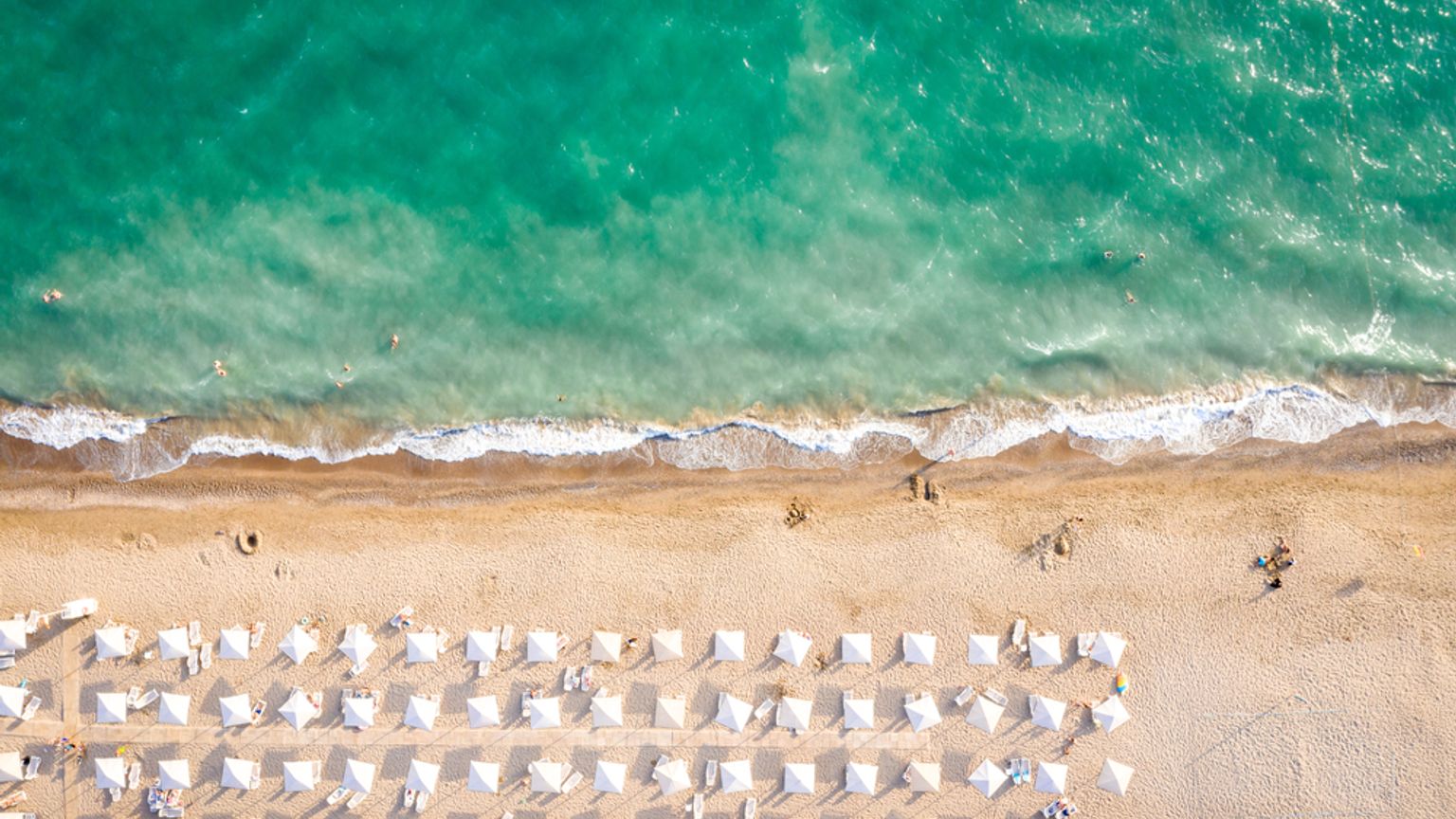 Vue aérienne d'une plage avec de l'eau turquoise, un rivage sablonneux et de nombreux parasols et transats blancs. Belek, Turquie