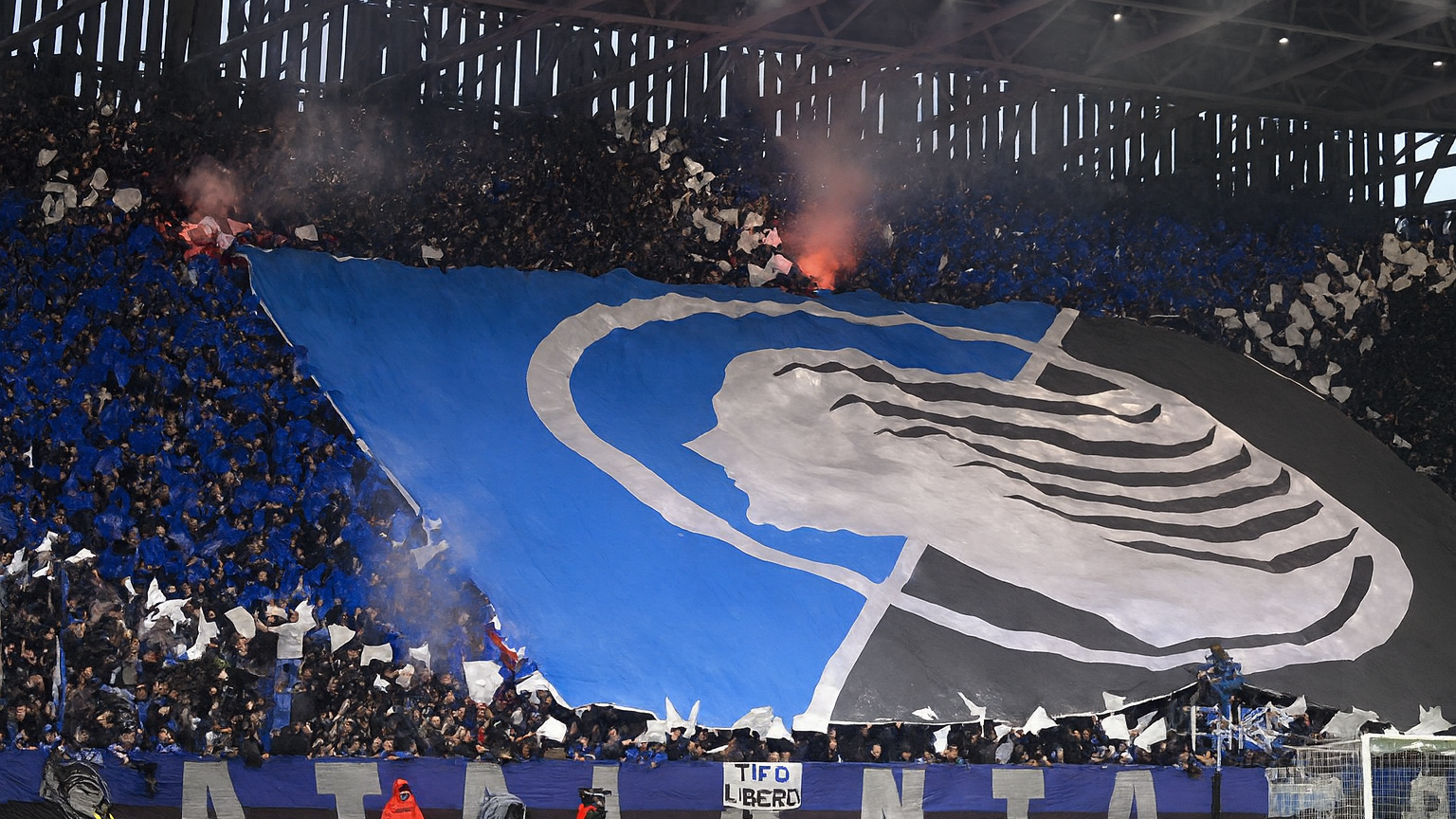 Football fans in a stadium holding a giant tifo with the Atalanta goddess logo and blue, black, and white colors, with flares smoking above.