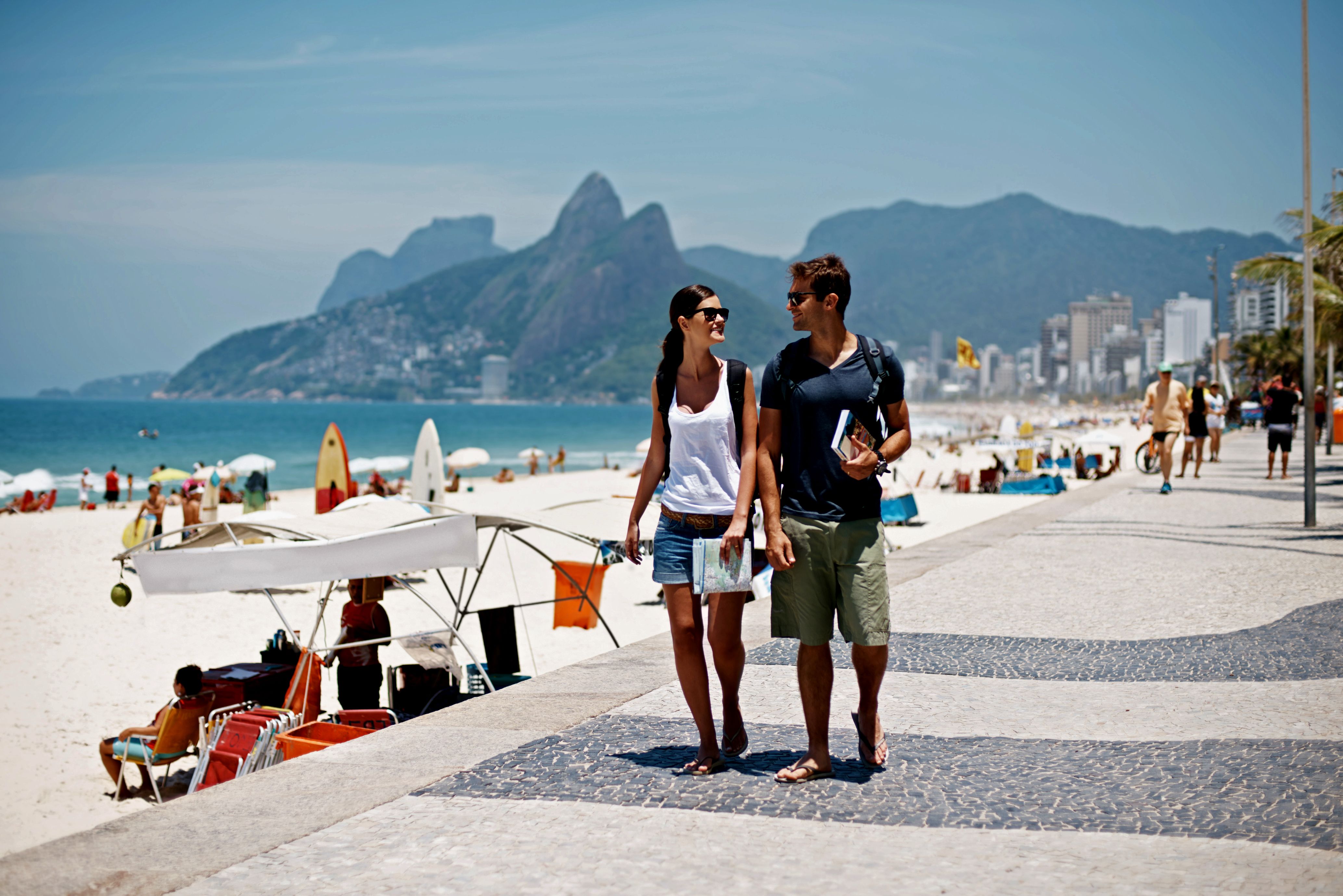 Ett par promenerar på en strandpromenad bredvid en solig strand, med berg och en stad i bakgrunden.