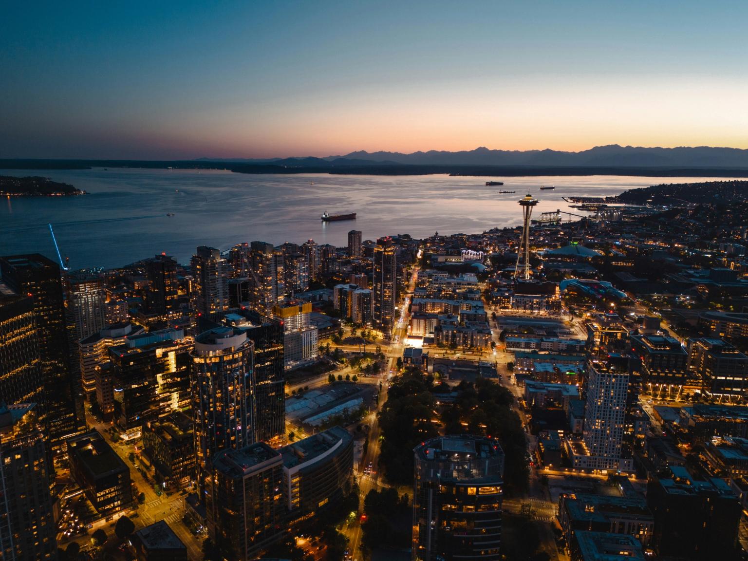 Luftfoto af Seattle i skumringen med Space Needle, oplyst bysilhuet, Puget Sound og fjerne bjerge.