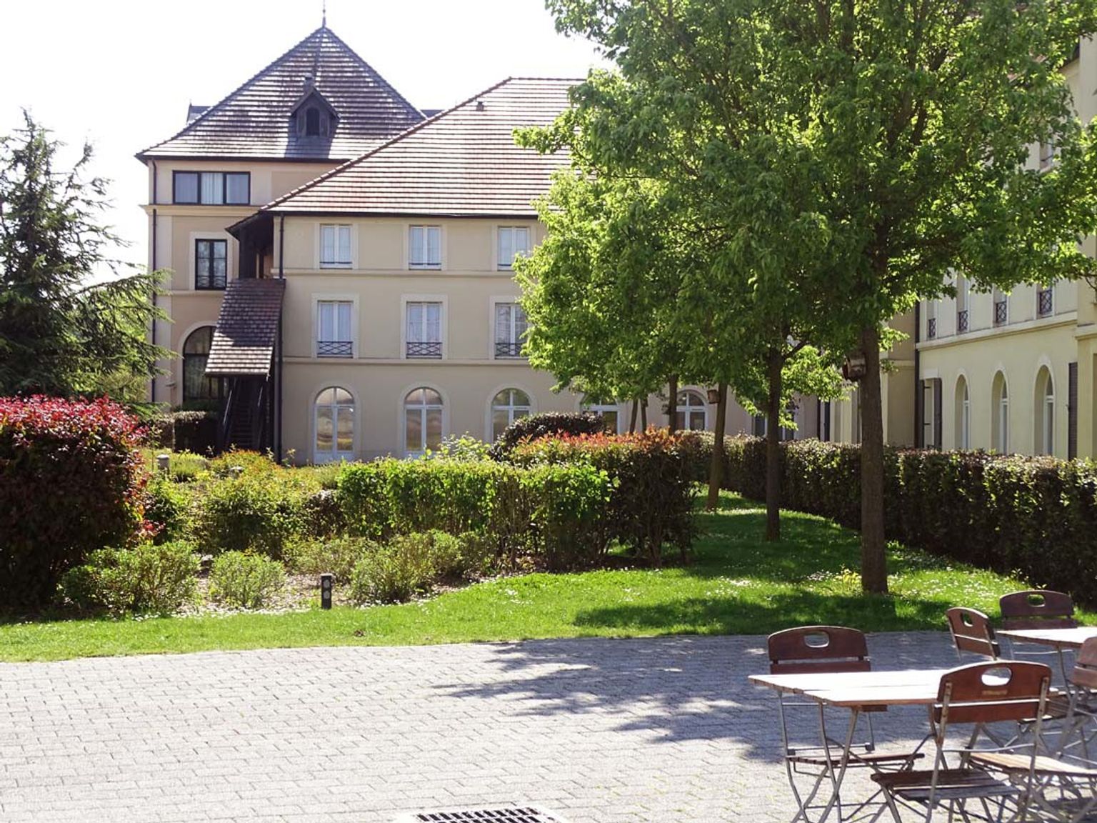 En stor byggnad med ett brunt tak, en grön trädgård och en stenlagd uteplats med utemöbler. Hotel Campanile Val de France vid Disneyland® Paris