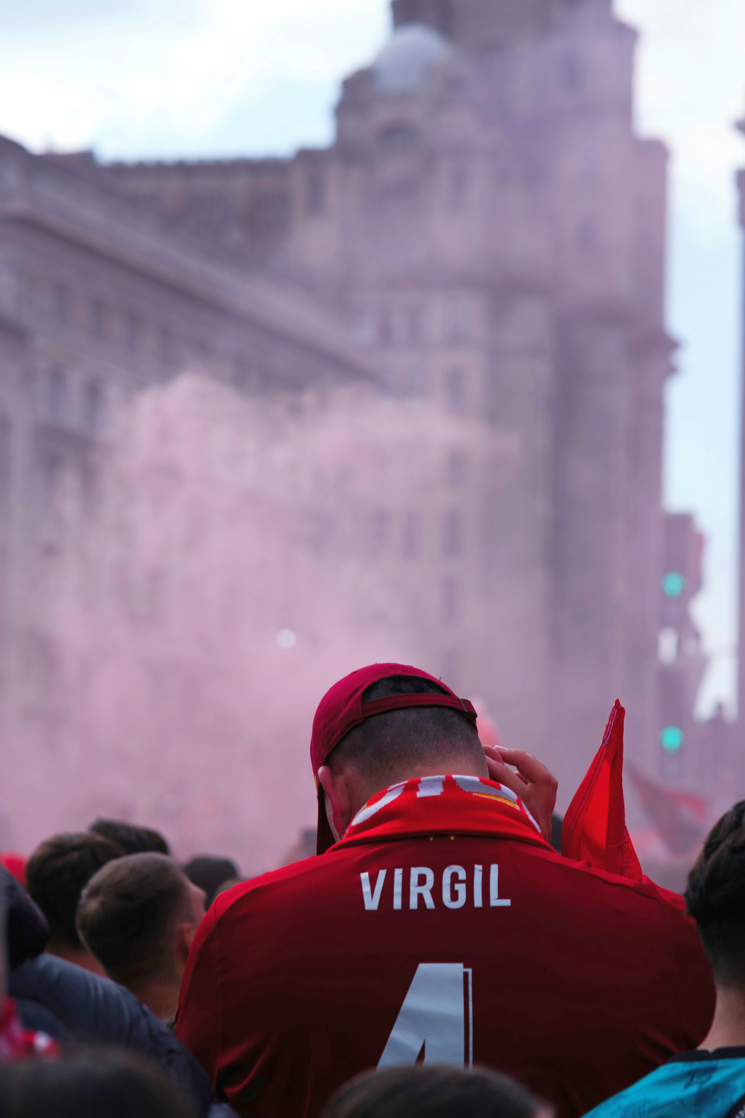 Fanin selkä punaisessa 'VIRGIL 4' pelipaidassa ja lippalakissa, vaaleanpunaisen savun ja sumeiden kaupunkirakennusten keskellä.