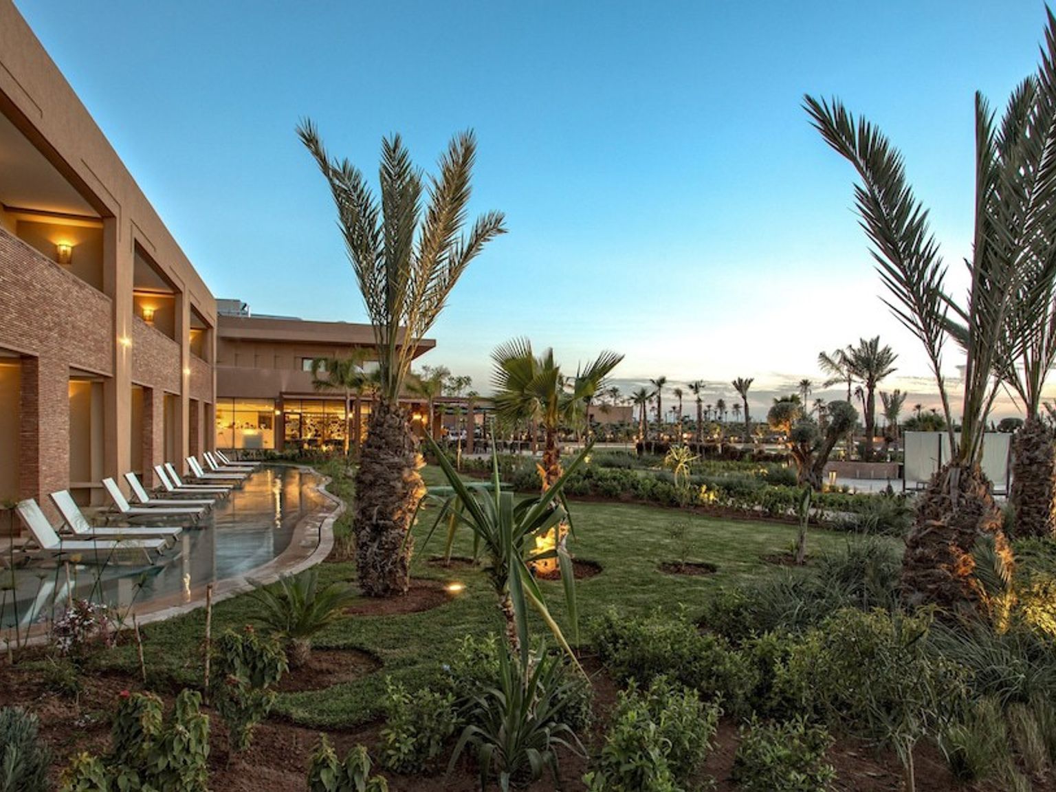 Kveldsutsikt over et feriested med en bygning, solsenger ved bassenget og palmefylte hager.