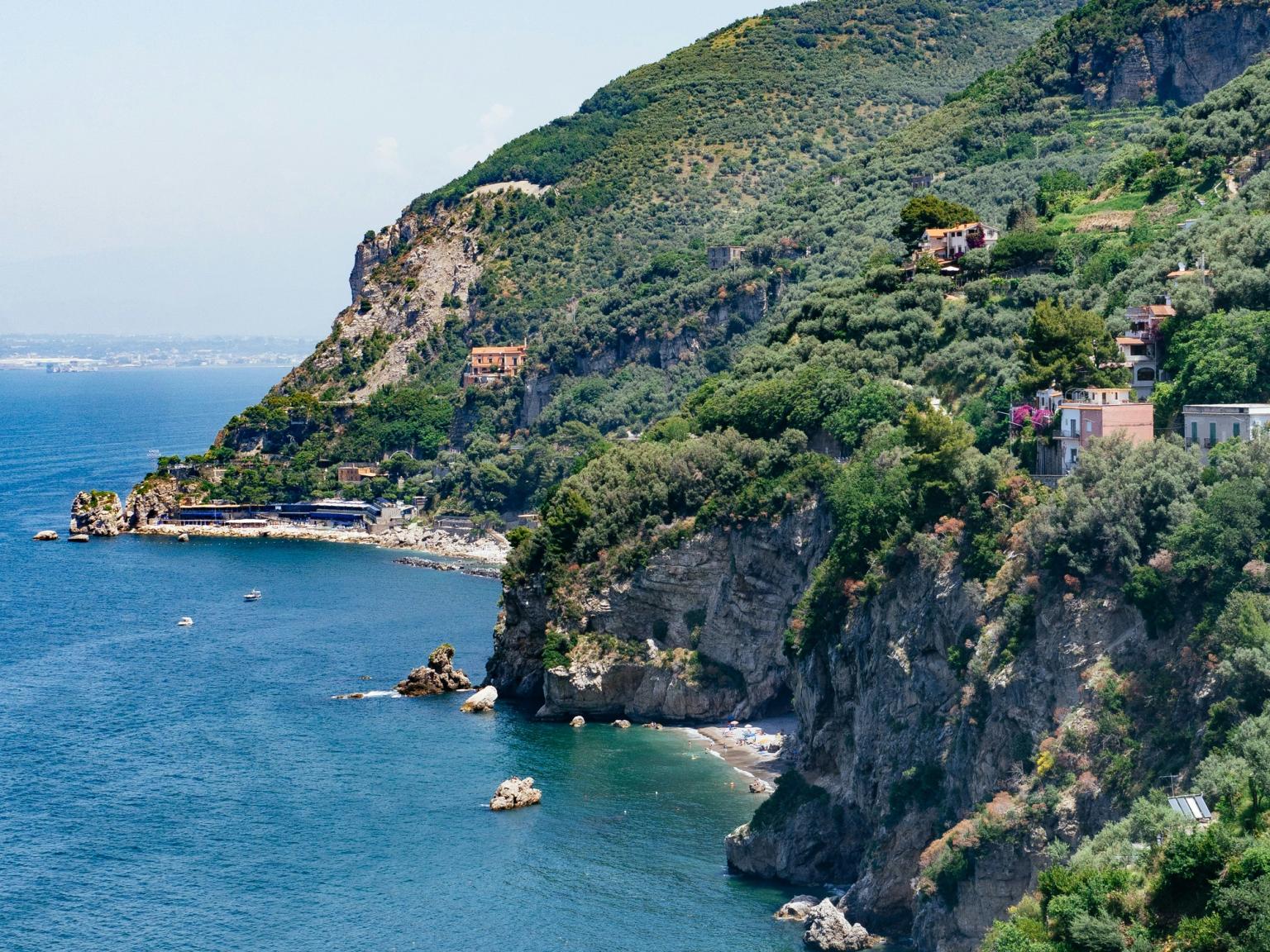 Vista costiera panoramica con ripide scogliere ricoperte di alberi che si affacciano su un mare blu profondo, punteggiato da piccole spiagge e case sulle colline. Vico Equense, Italia