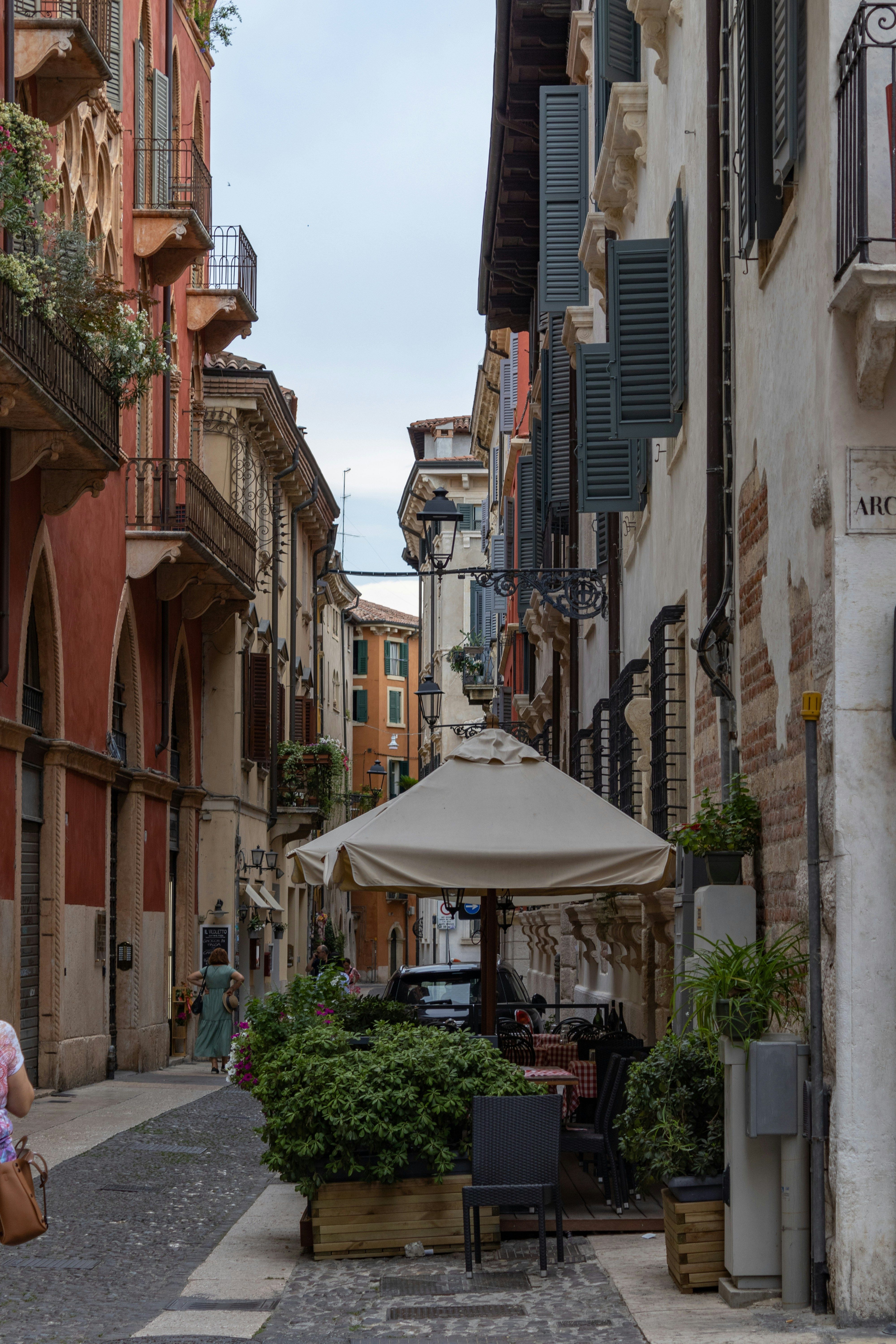 En smal brosteinsgate i en historisk europeisk by, flankert av fleretasjes bygninger med balkonger og skodder, og en utendørskafé med en stor parasoll.