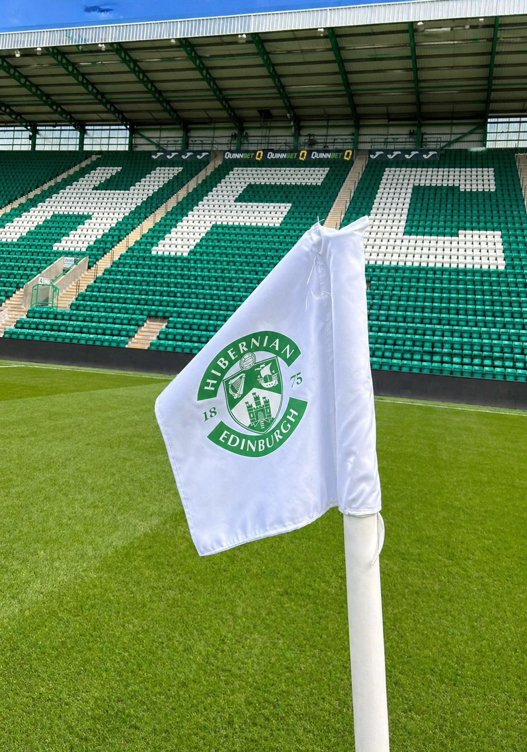 A white Hibernian FC corner flag on a green football pitch with stadium seats in the background spelling "HIFC".