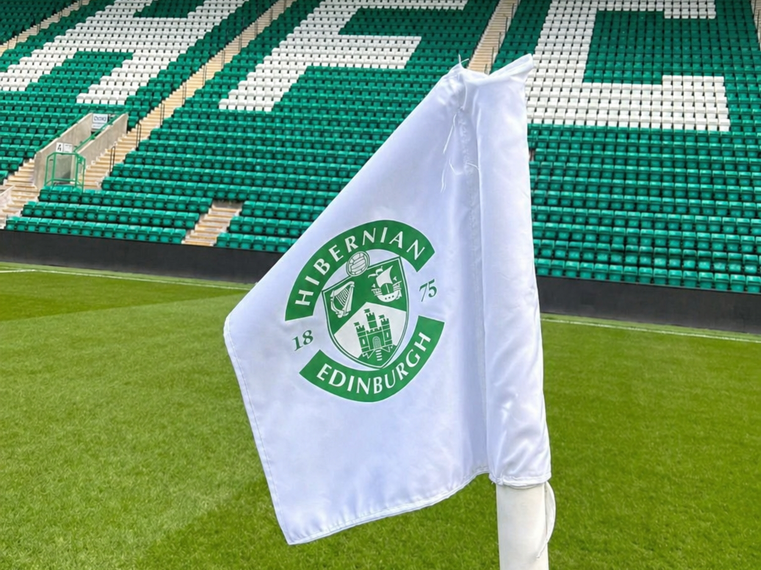 A white Hibernian FC corner flag on a green football pitch with stadium seats in the background spelling "HIFC".