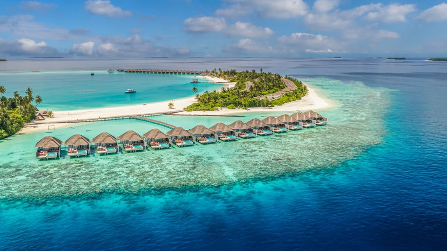 Vista aérea de un complejo turístico en una isla tropical con bungalós sobre el agua, playa de arena blanca y aguas turquesas cristalinas.