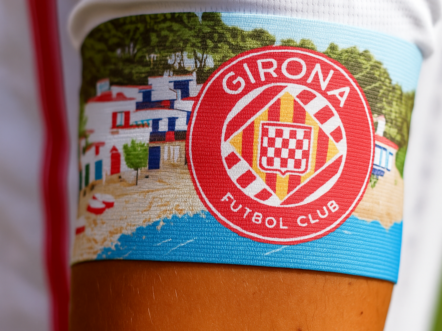 Kolorowa opaska na ręce, przedstawiająca scenę miasteczka plażowego i czerwone logo klubu piłkarskiego Girona.