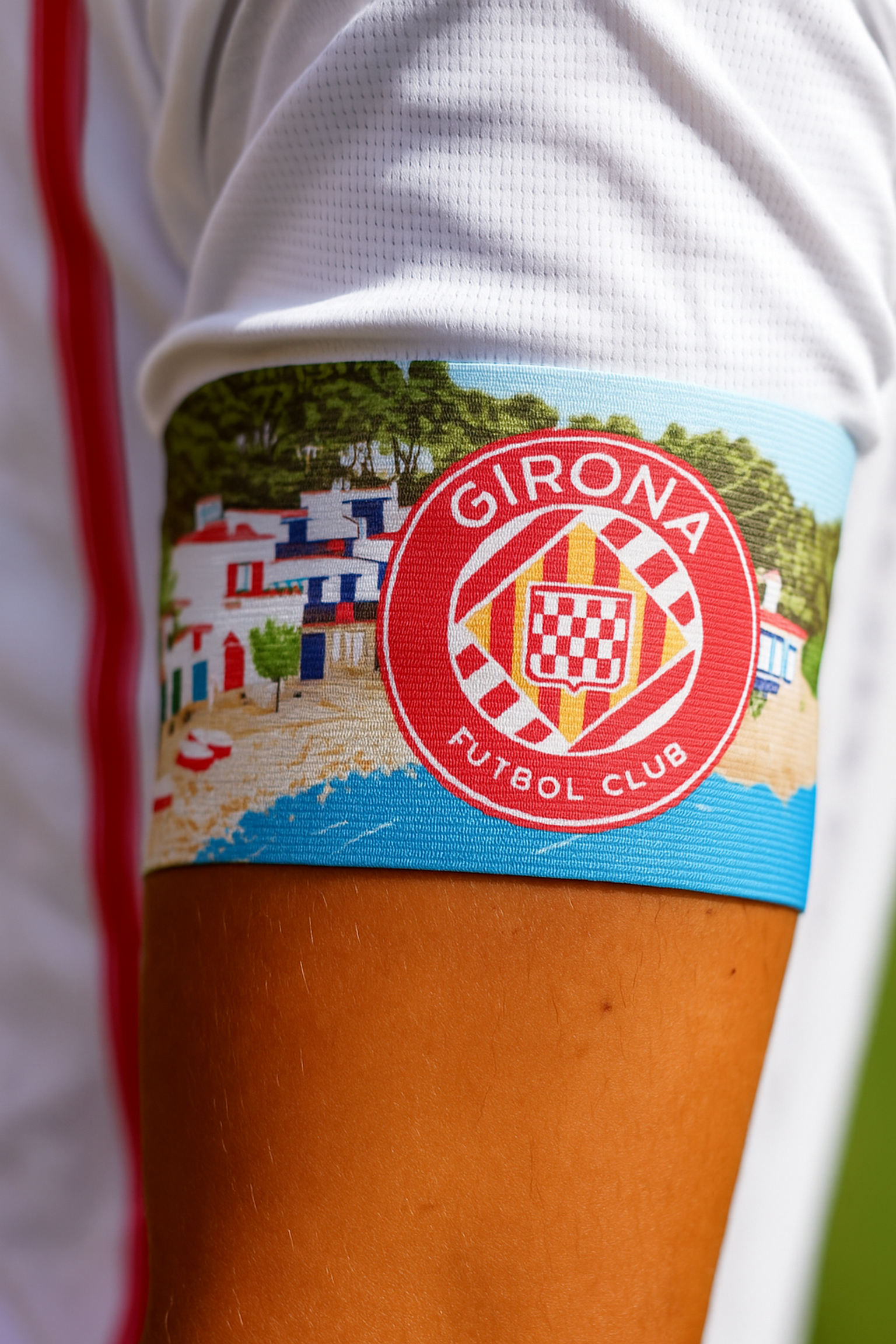 Kolorowa opaska na ręce, przedstawiająca scenę miasteczka plażowego i czerwone logo klubu piłkarskiego Girona.