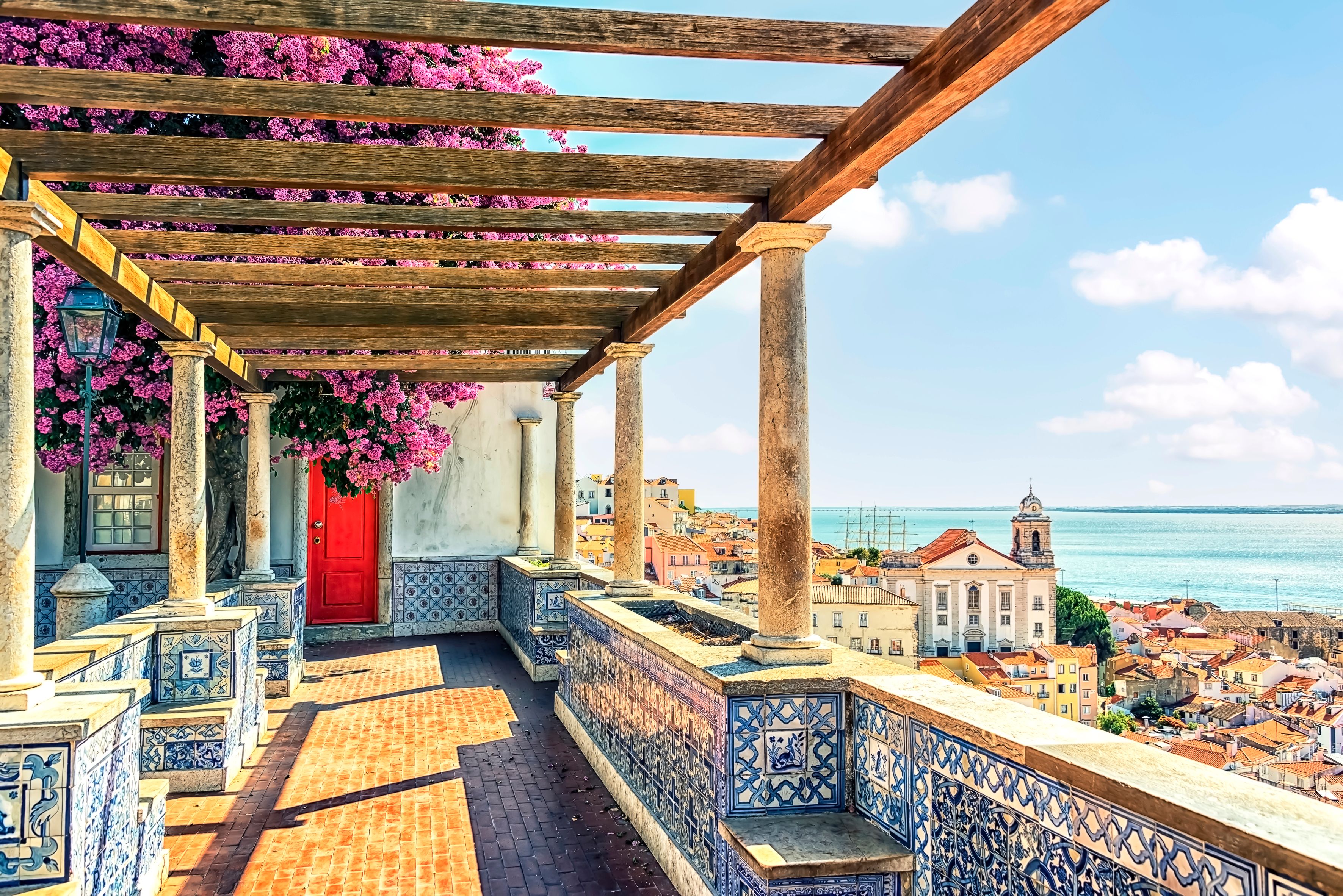 Terrasse med en pergola dækket af lyserød bougainvillea og blå azulejofliser med udsigt over en kystby og havet.