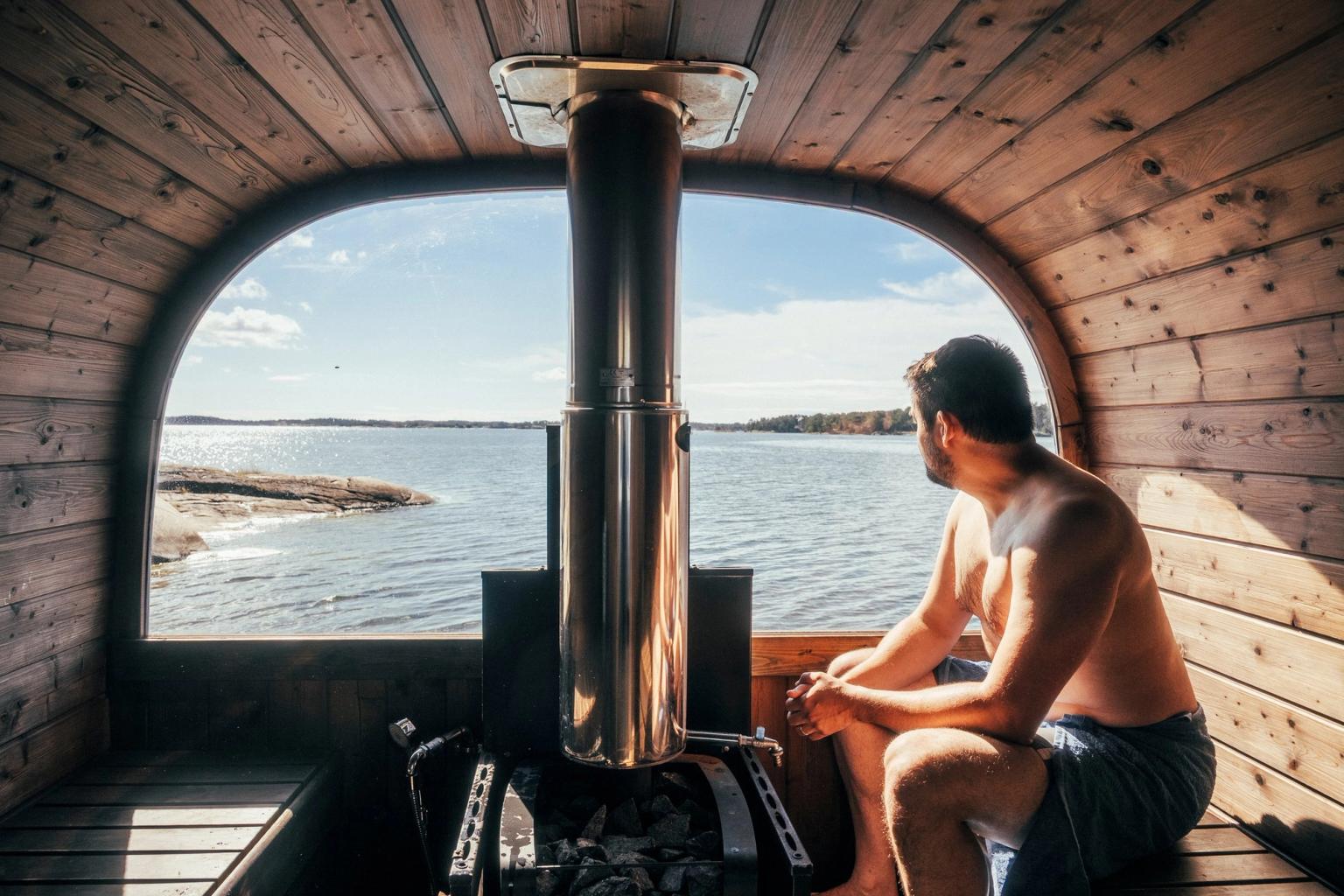 En mand sidder i en tøndesauna af træ og kigger ud gennem et buet vindue mod en sø med stenet kyst.