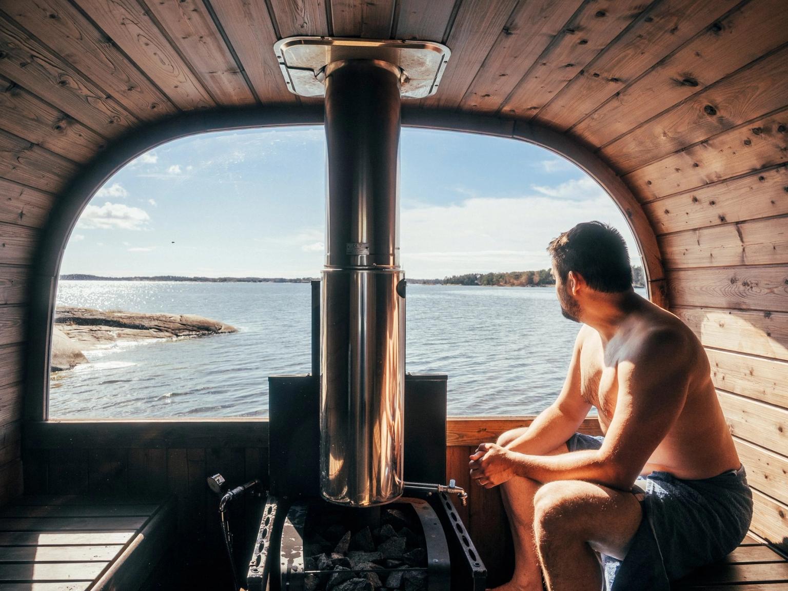En mand sidder i en tøndesauna af træ og kigger ud gennem et buet vindue mod en sø med stenet kyst.
