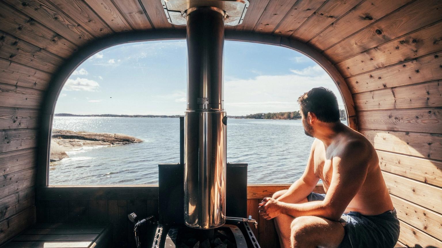 En mand sidder i en tøndesauna af træ og kigger ud gennem et buet vindue mod en sø med stenet kyst.