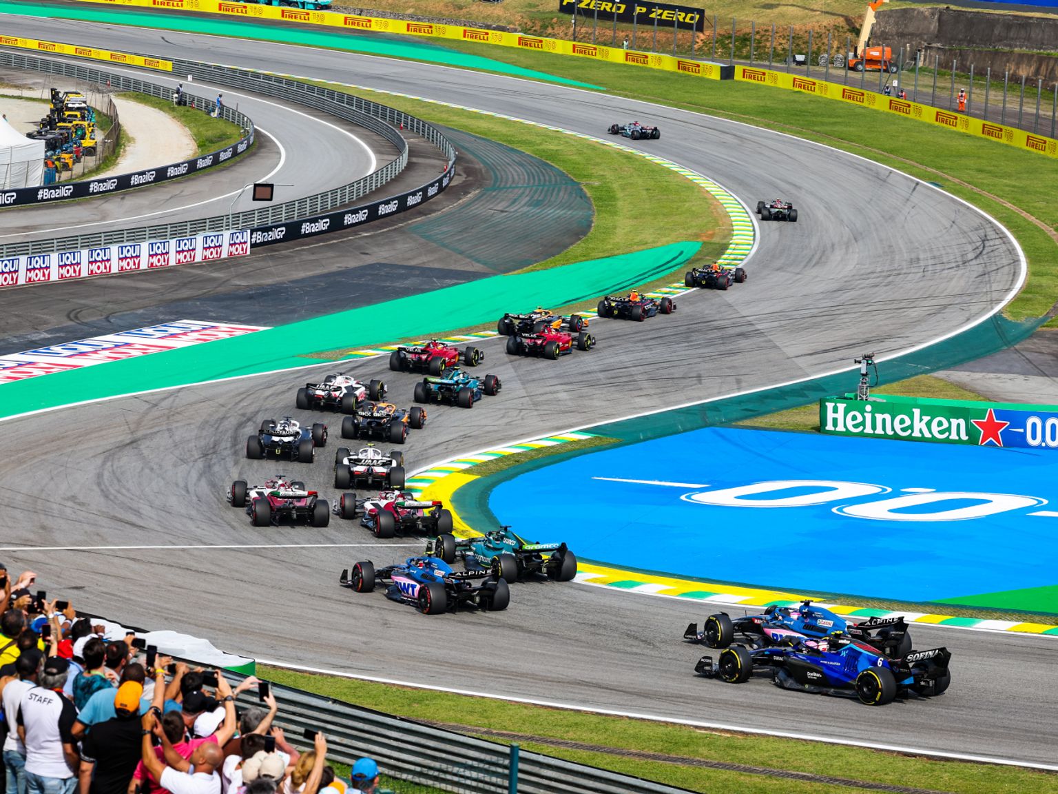 F1 cars race around a corner on a track, with spectators in the foreground.