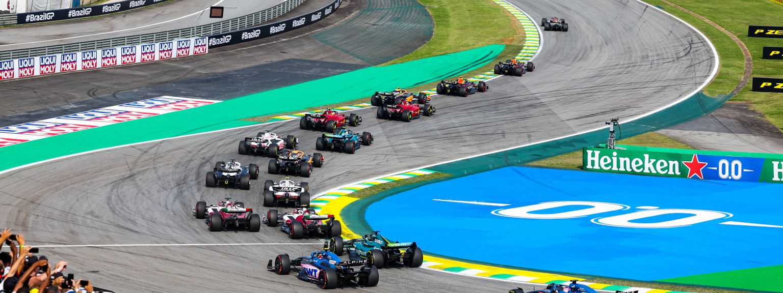 F1 cars race around a corner on a track, with spectators in the foreground.