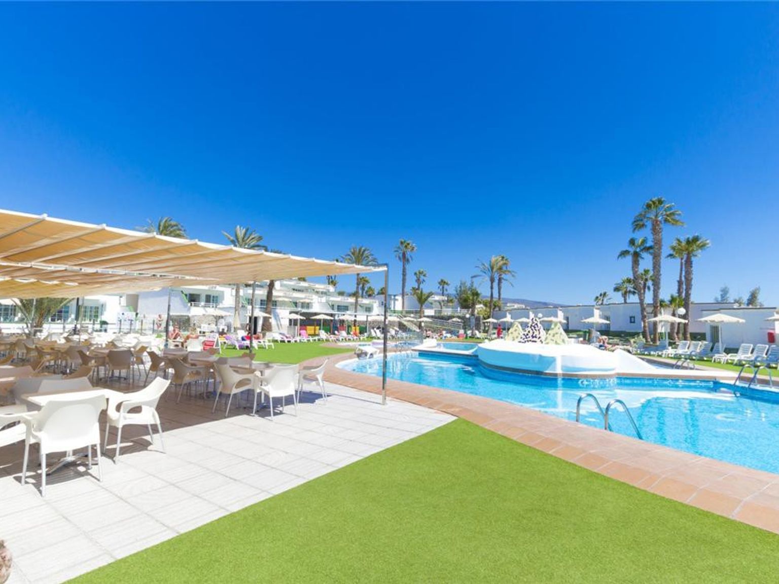 Solrikt feriested med en utendørs spiseplass, svømmebasseng og palmer under en klar blå himmel.