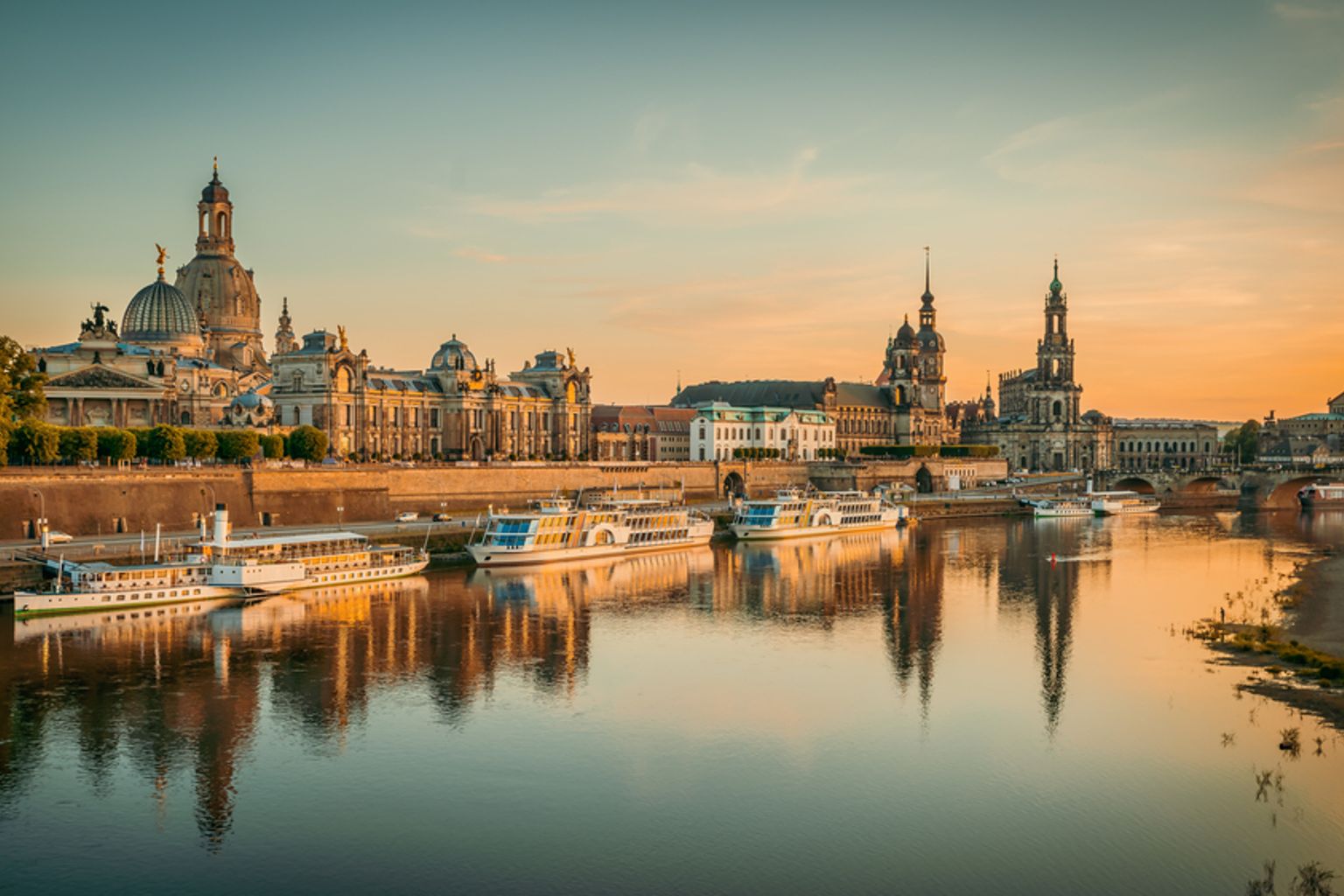Dresdens Stadtsilhouette bei Sonnenaufgang, die historische Gebäude und Boote zeigt, die sich im ruhigen Elbe-Fluss spiegeln.