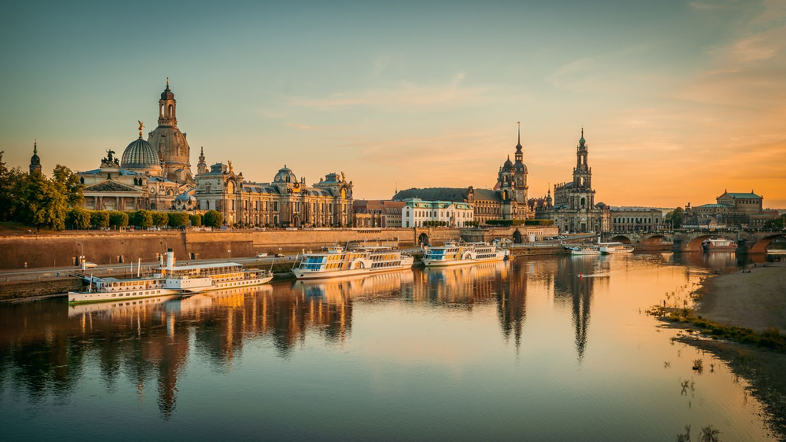 Dresdens Stadtsilhouette bei Sonnenaufgang, die historische Gebäude und Boote zeigt, die sich im ruhigen Elbe-Fluss spiegeln.