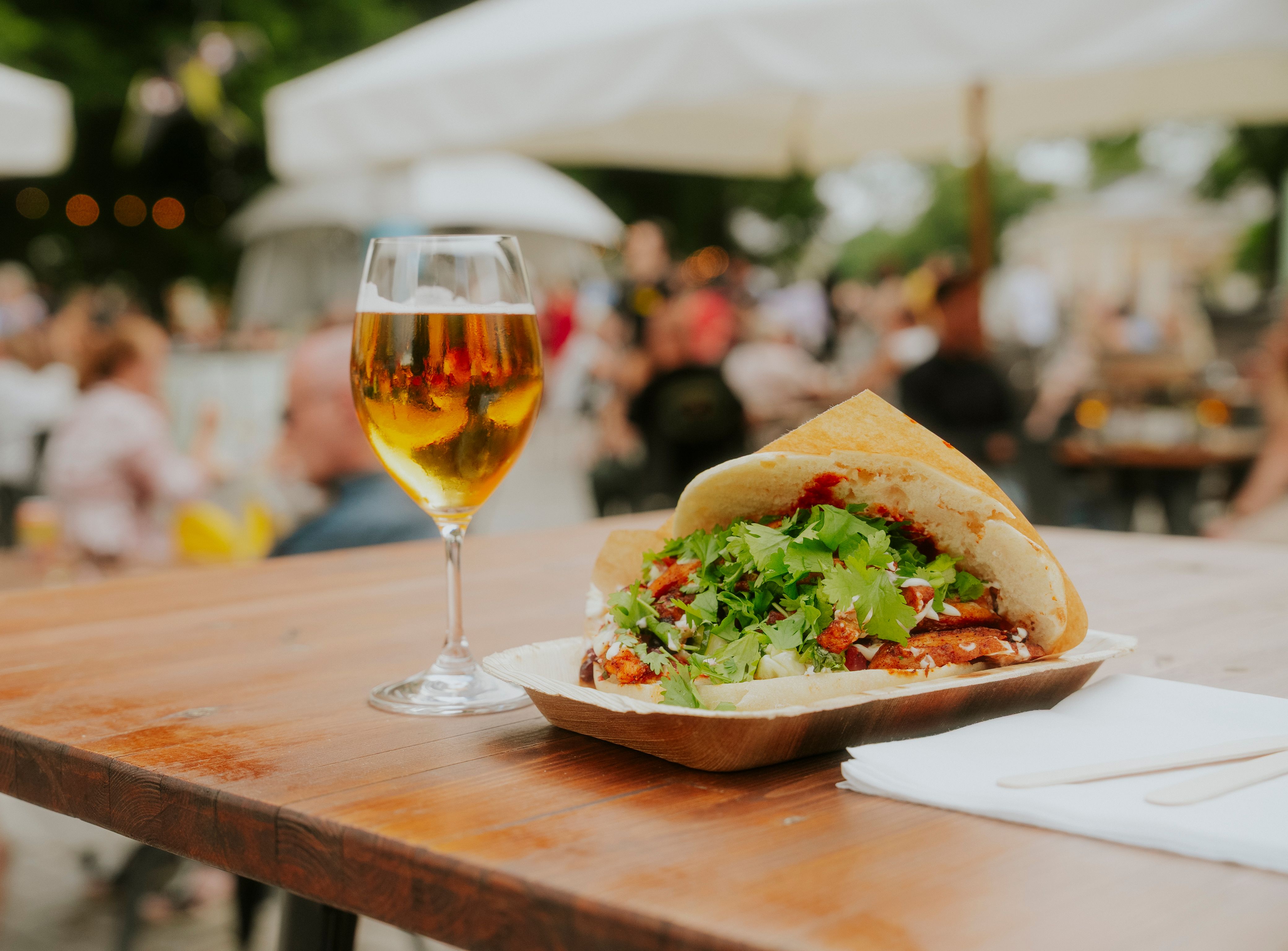 Et glas øl ved siden af en kebab i fladbrød på et træbord ved et udendørs arrangement.
