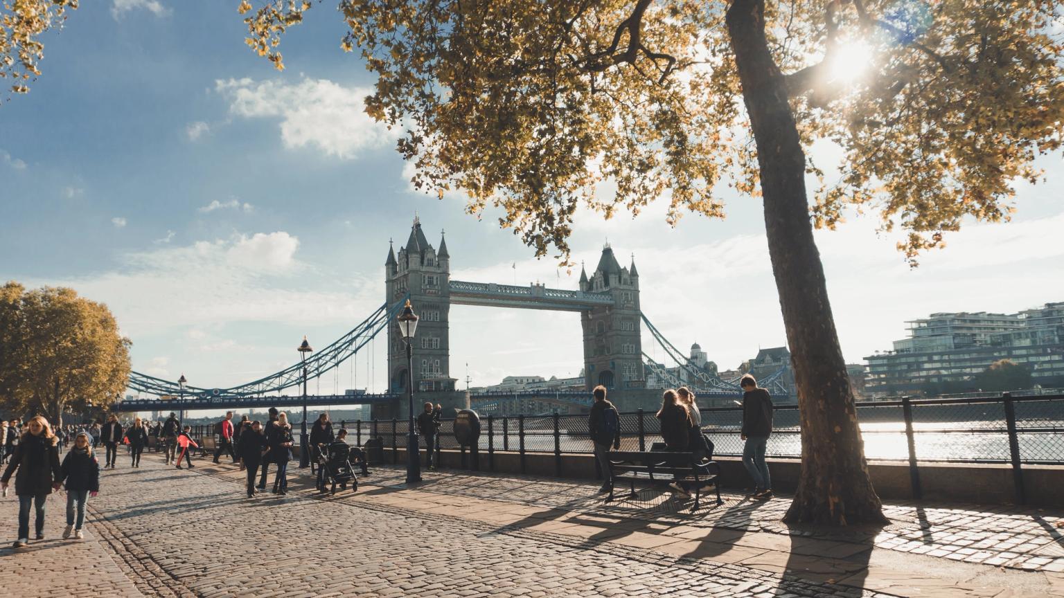 a group of people are walking along a cobblestone sidewalk in front of the tower bridge in london