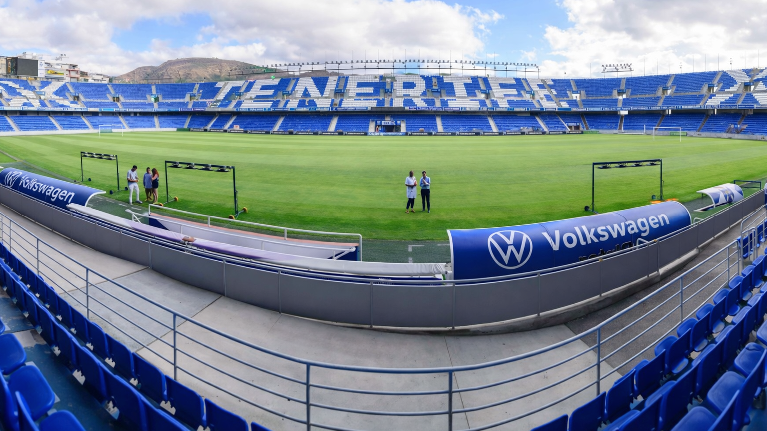 Un estadio de fútbol vacío azul y verde con "TENERIFE" en las gradas y un anuncio de "Volkswagen" en la línea lateral.
