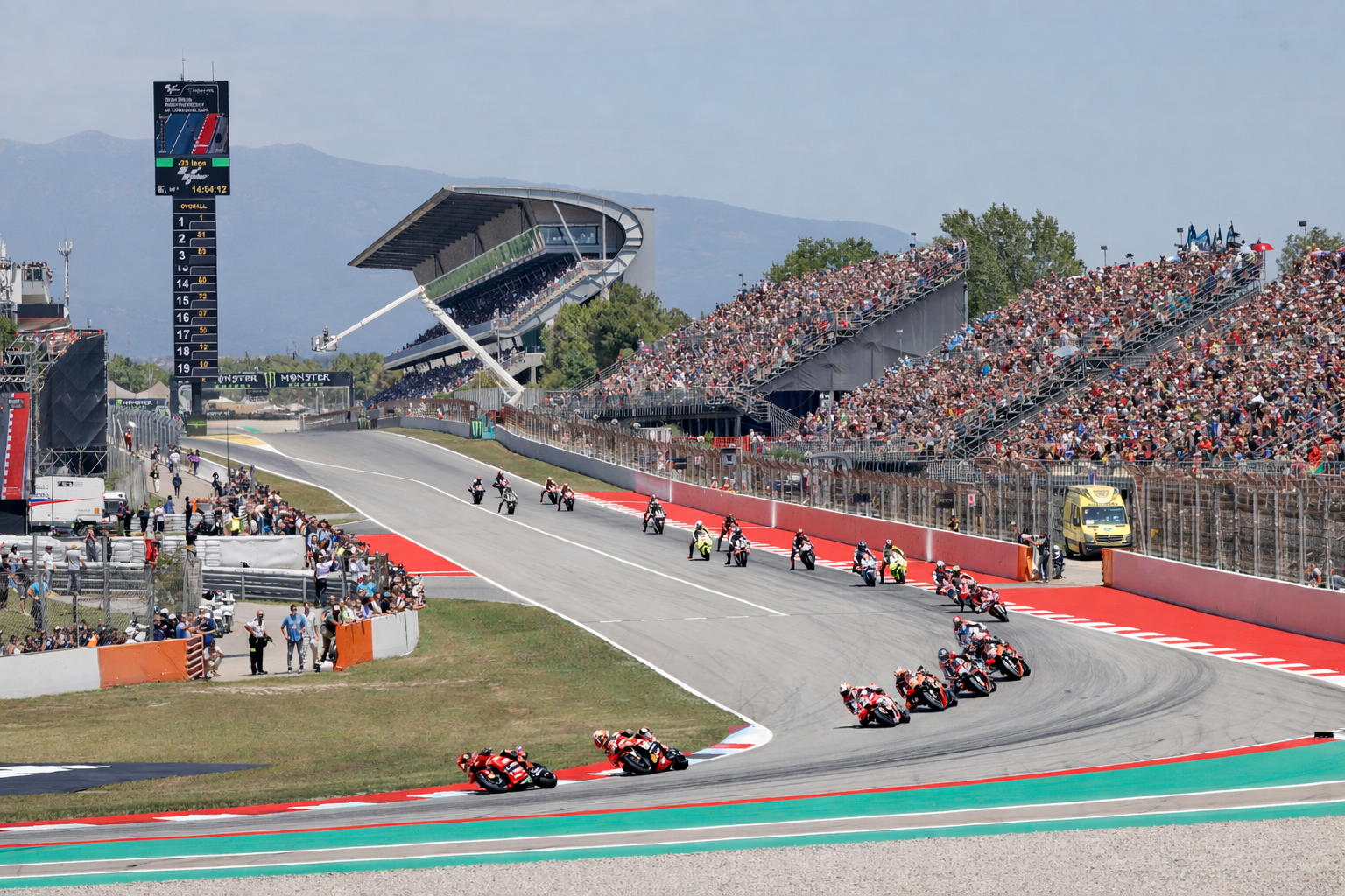 Motorcycles race around a bend on a track, with crowded grandstands under a sunny sky.