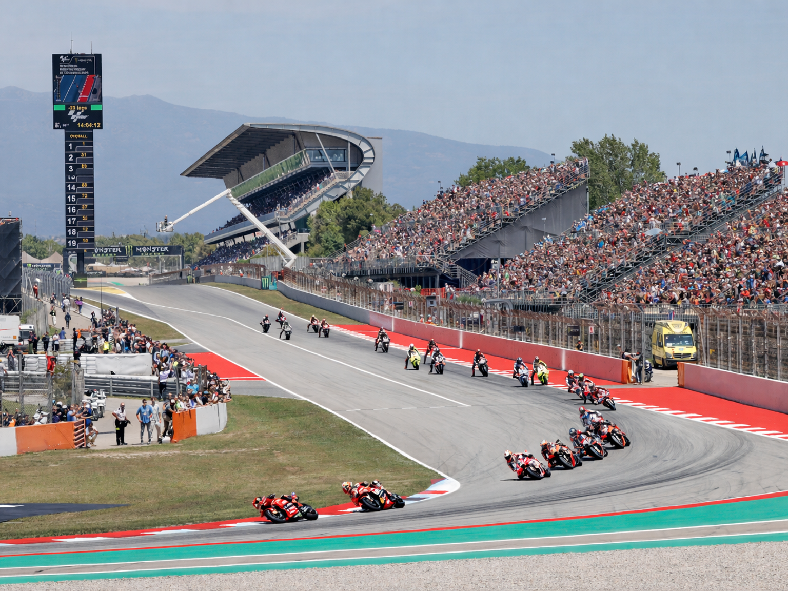 Motorcykler kører ræs rundt i et sving på en bane, med fyldte tribuner under en solrig himmel.
